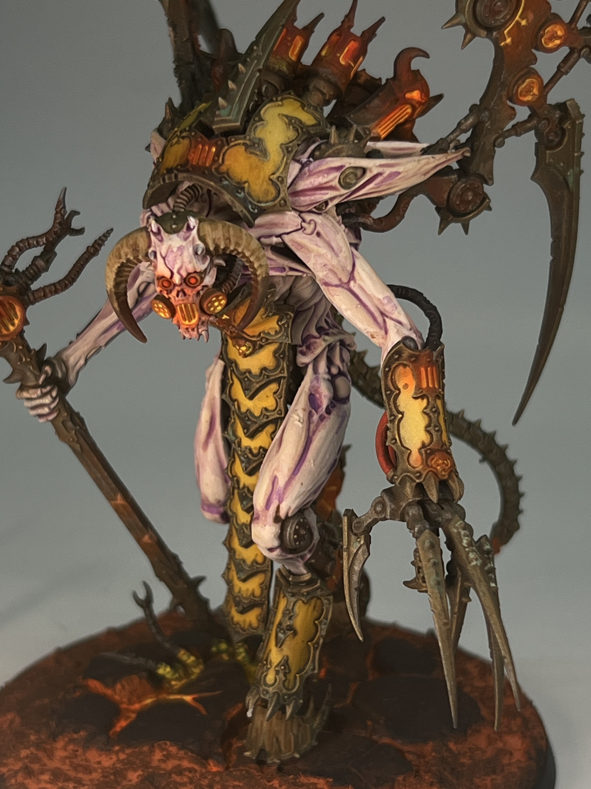 A detailed miniature figurine of a fantasy creature with a skull face, ram horns, and a long tongue, holding a staff, wearing armor, with glowing orange accents, and large wings, standing on a base resembling molten lava.