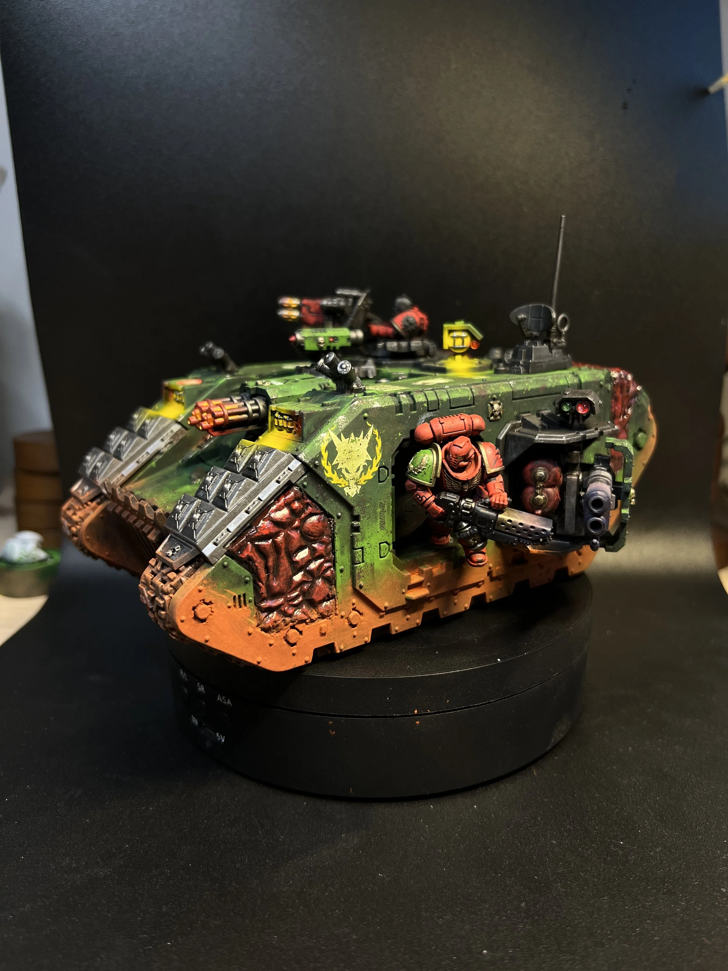 A miniature tank model painted in green with red and black accents, featuring a yellow emblem on its side and a soldier figure with red armor and a gun, placed on a circular black base.