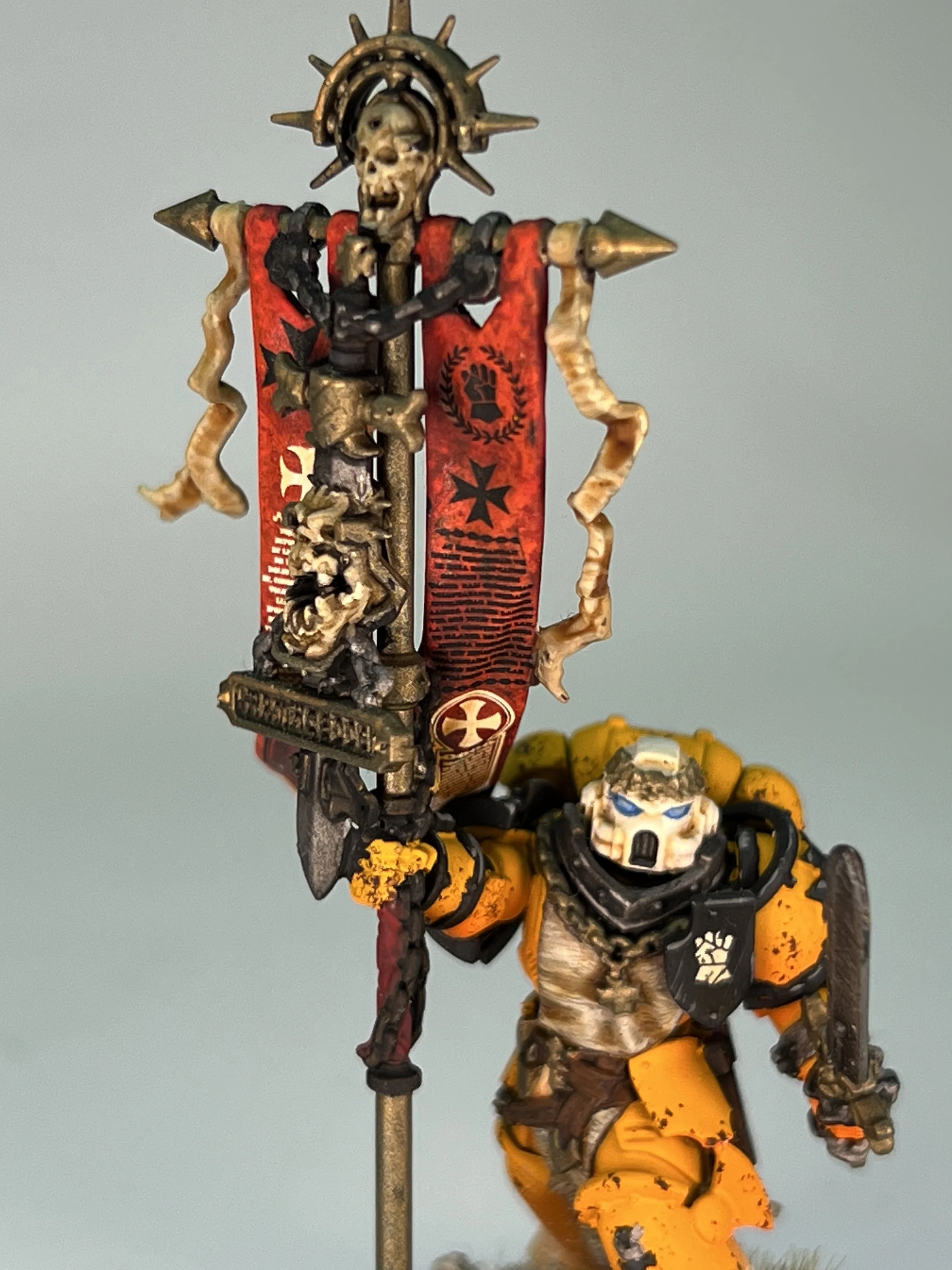 Close-up of a detailed miniature figurine, likely from a tabletop game, featuring a skull-faced character in armor holding a spear with a skull at the top. The character has a red banner with black symbols and a skull emblem, and is dressed in yellow