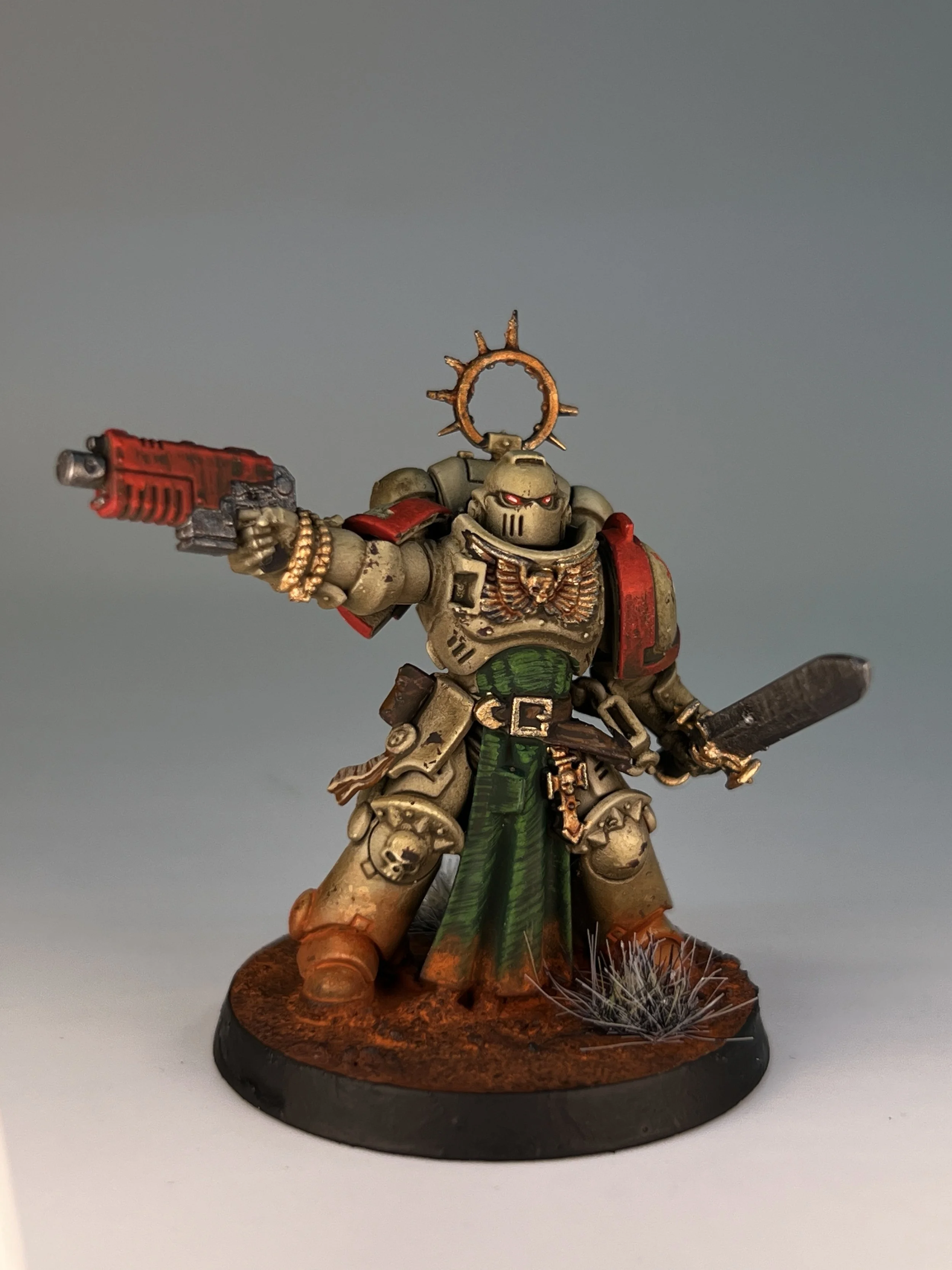 A detailed miniature figurine of a sci-fi soldier or warrior, painted in beige, red, and green, standing on a textured base with grass tufts, holding a gun, with a minimal background.