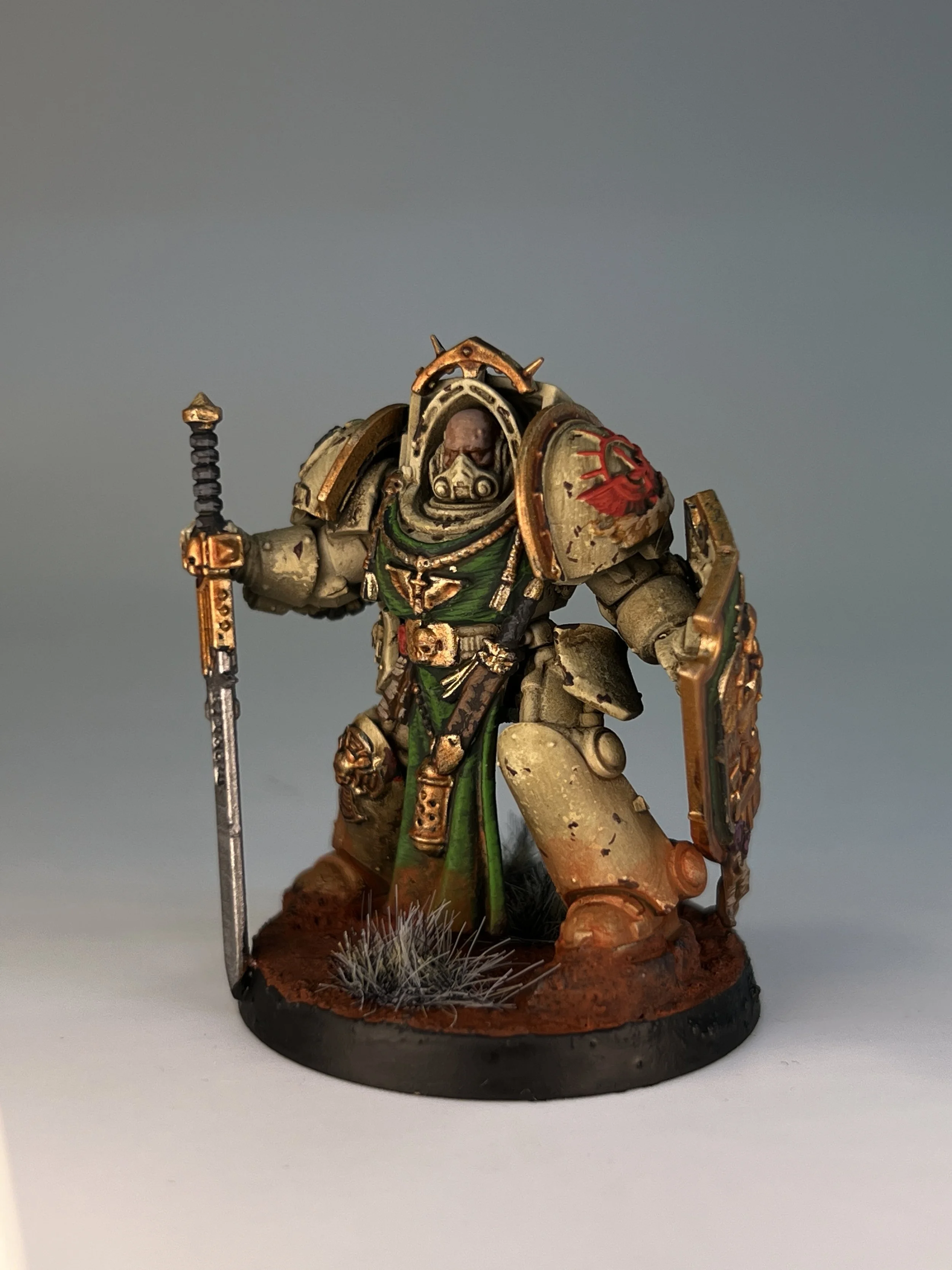 A detailed miniature figure of a fantasy warrior in armor holding a sword and shield, standing on a round base with rocks and grass. The armor is painted in beige with gold and green accents, with a red emblem on the shoulder.