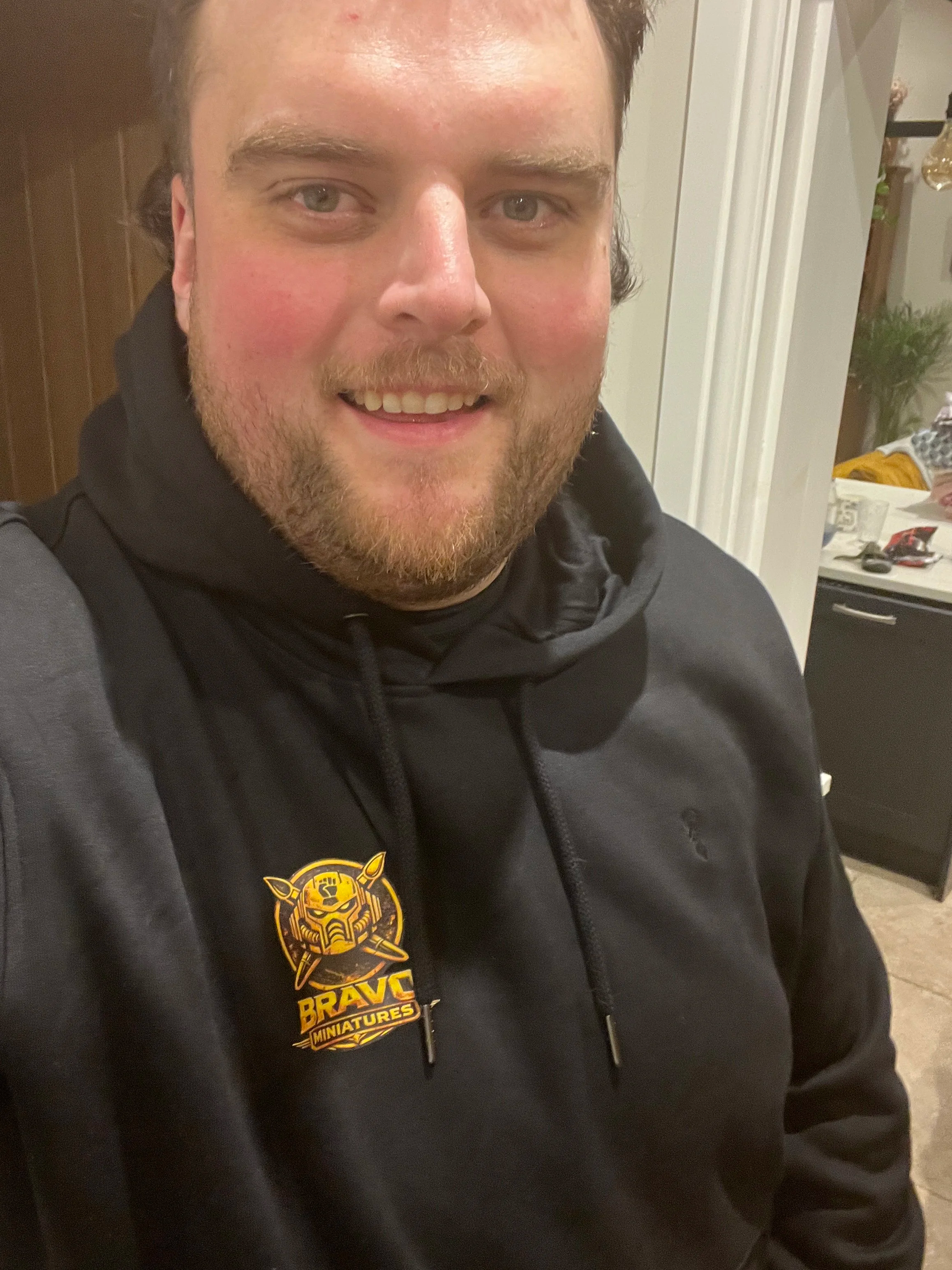 Close-up of a smiling man wearing a black hoodie with a 'Braves Miniatures' logo on it, in an indoor setting with a kitchen in the background.