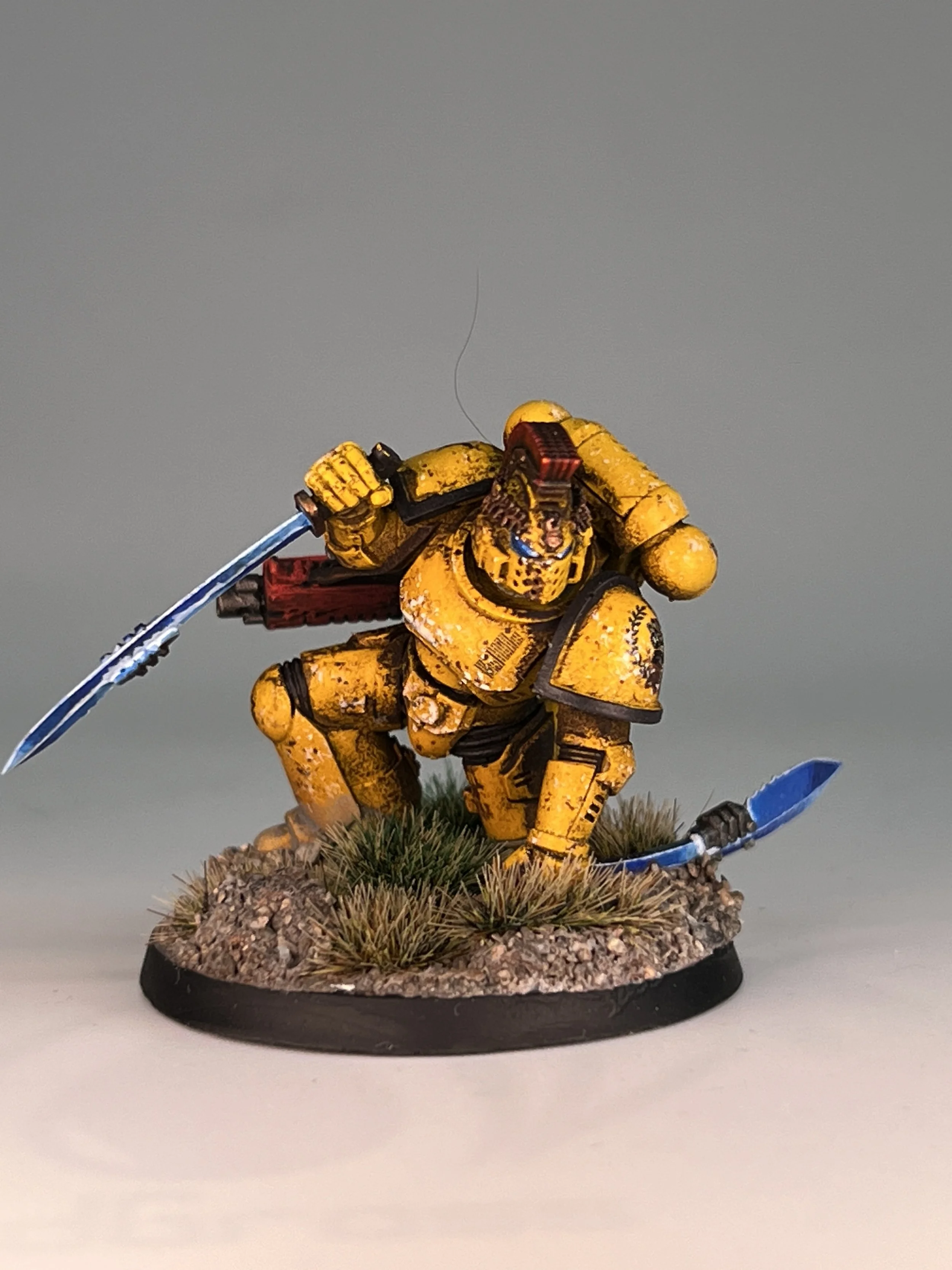 A miniature painted figurine of a soldier in yellow armor, holding a sword, standing on a round base with grass and rocks, against a plain gray background.
