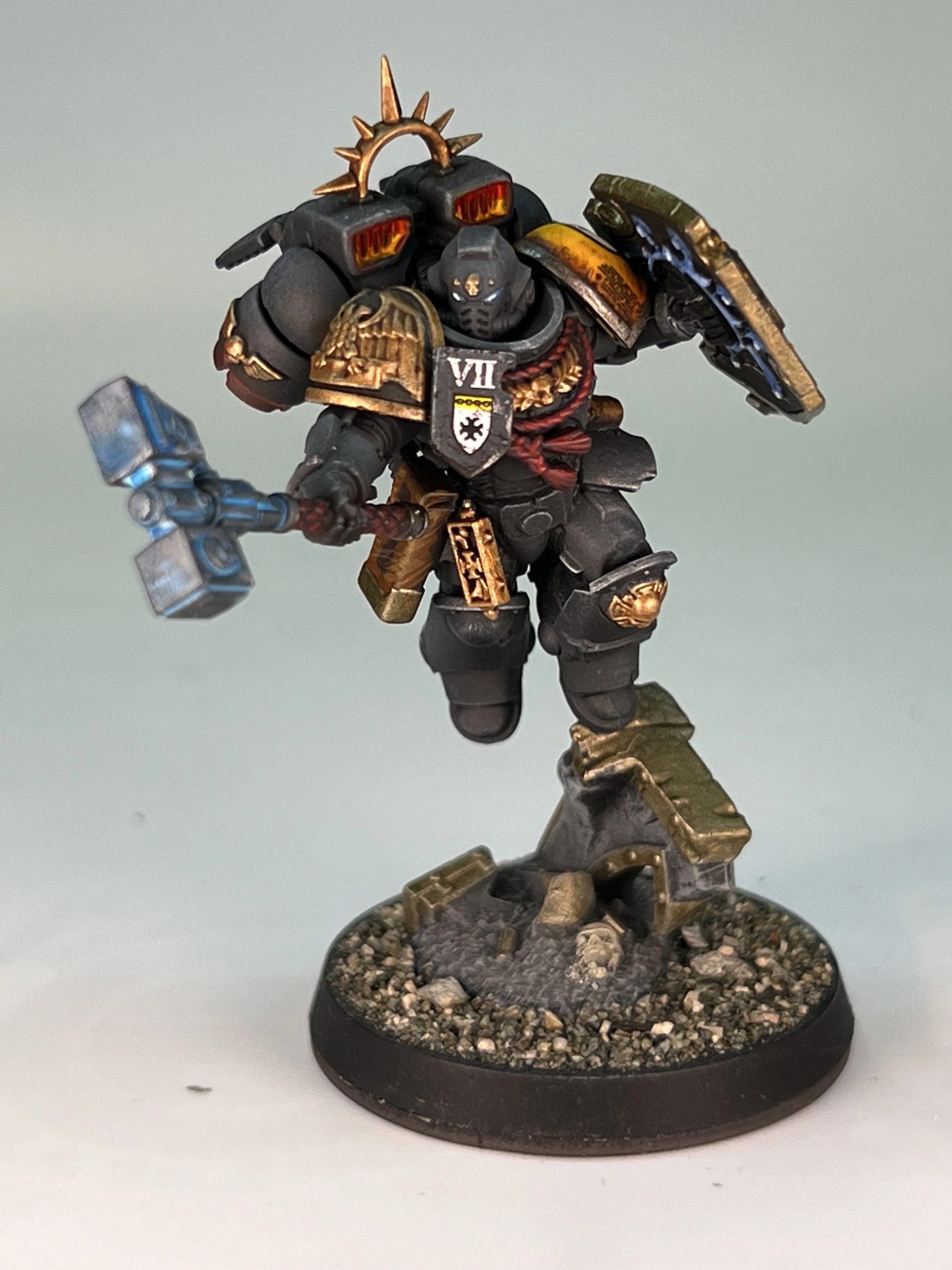 Miniature model of a sci-fi soldier in dark armor with gold accents, holding a hammer, standing on a textured round base.