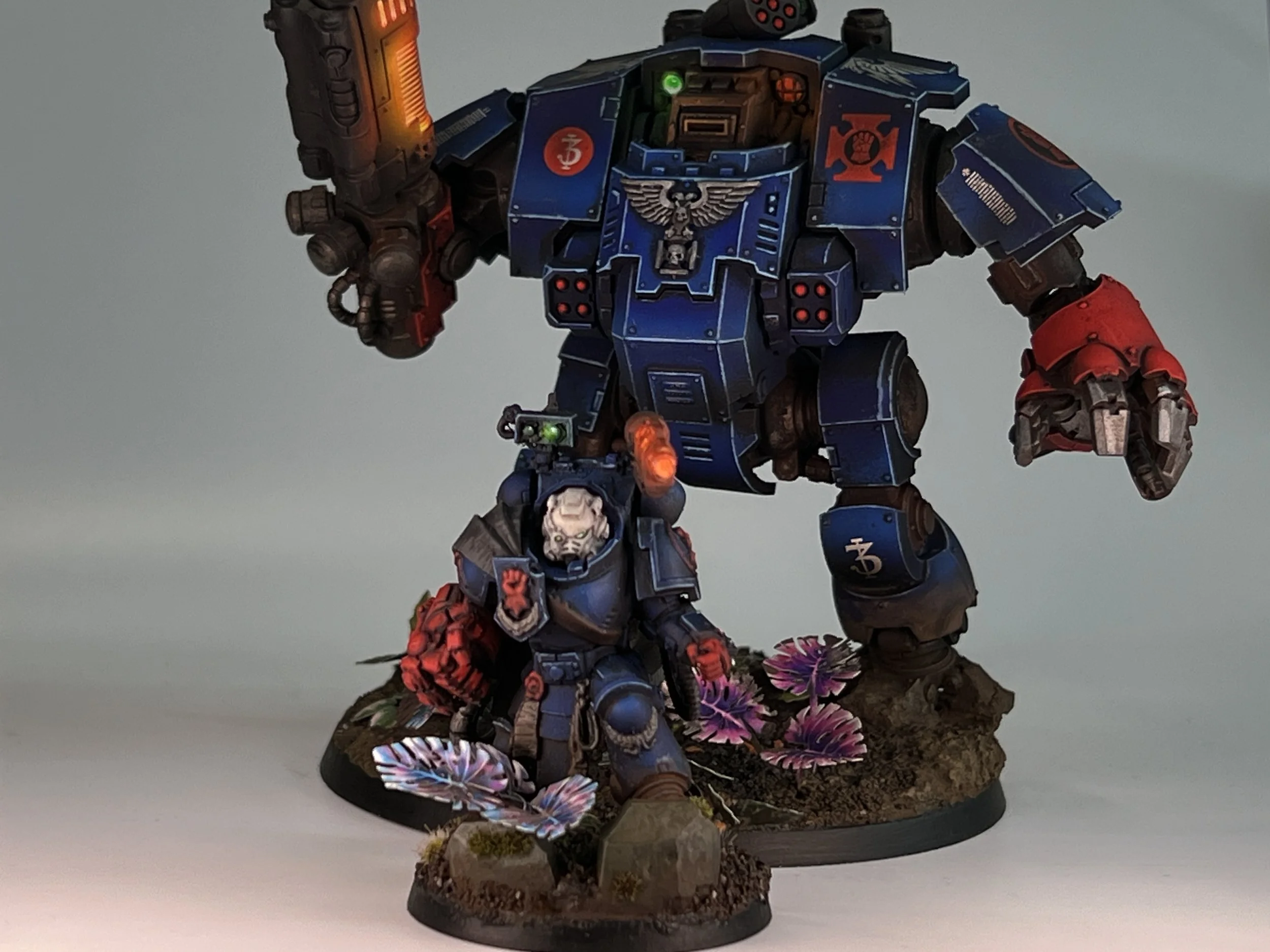 A detailed painted miniature of a war robot wearing blue armor with red accents, standing on a base decorated with flowers and terrain.