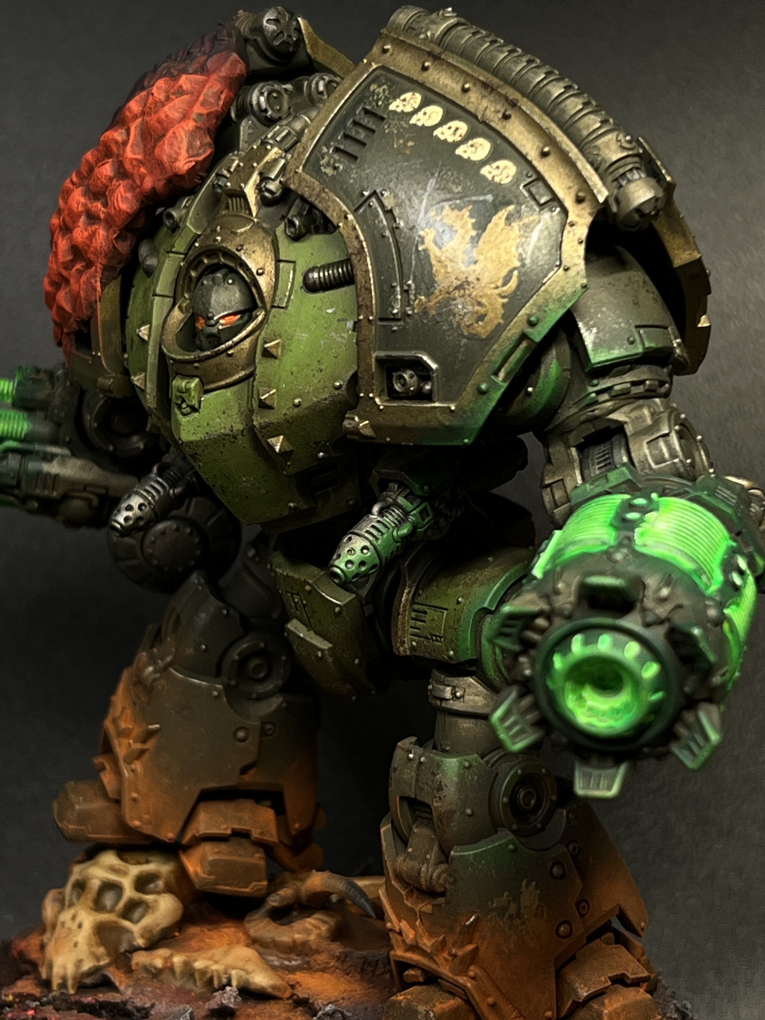A detailed figurine of a heavily armored sci-fi robot with glowing green elements, weathered green and metallic armor, and various mechanical and armor features, mounted on a rocky base with skulls and bones.