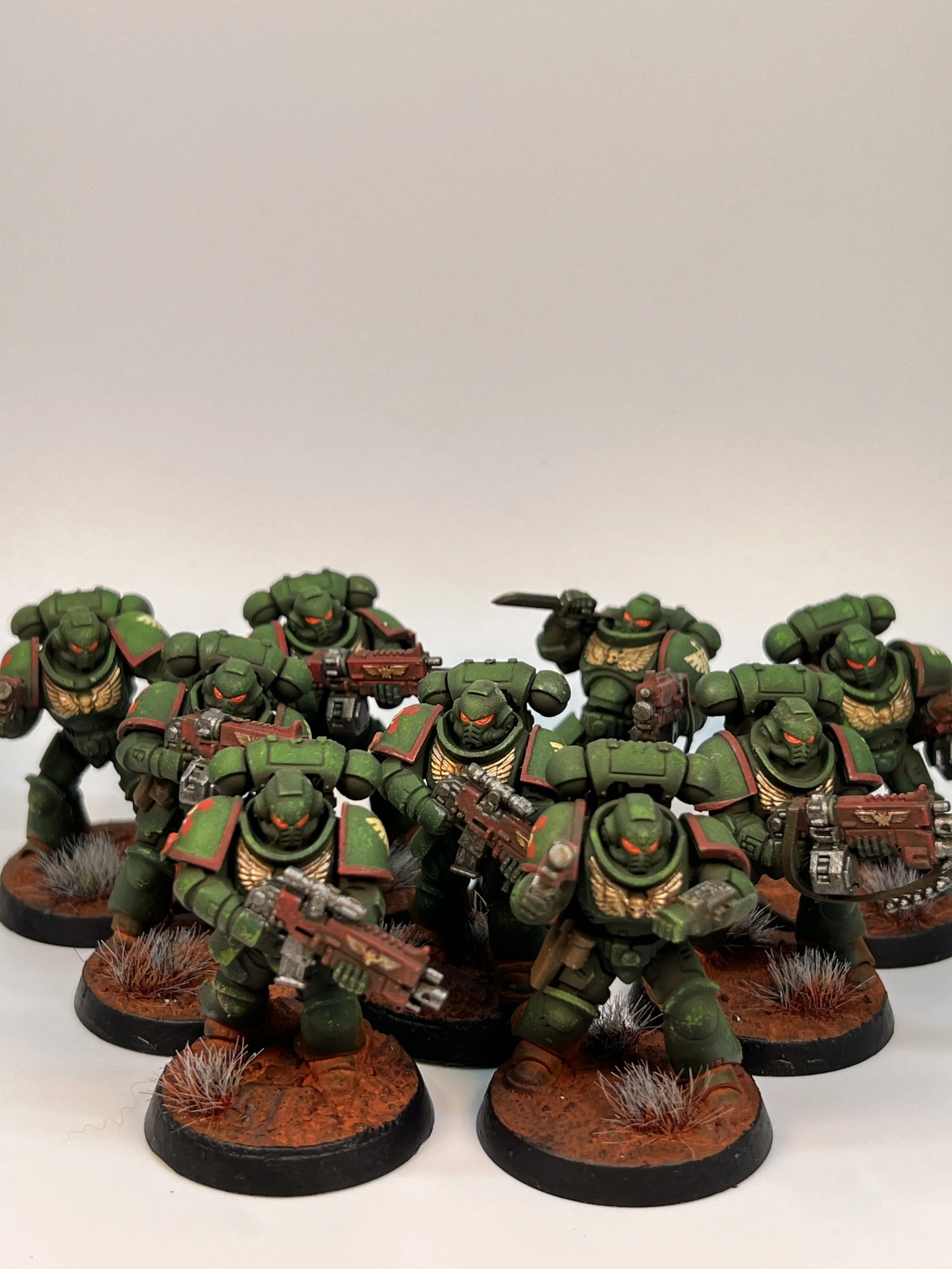 A group of miniature green armored soldiers with red eyes, holding guns, standing on round bases with terrain and tufts of grass in a gaming setup.