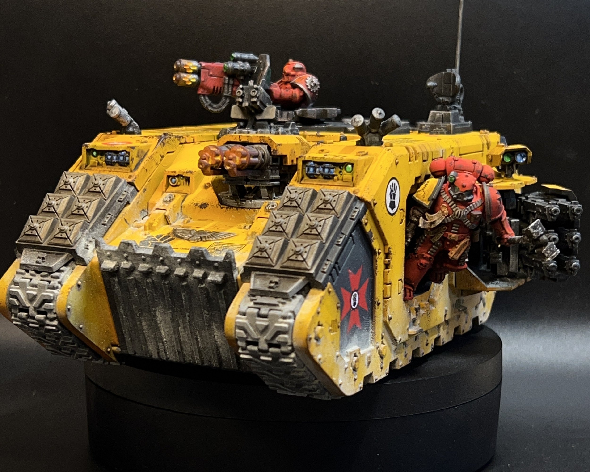 A detailed model of a yellow futuristic tank with a red armored soldier figure standing next to it. The tank features black tread tracks, a mounted gun on top, and various small guns and mechanical parts. The background is black.