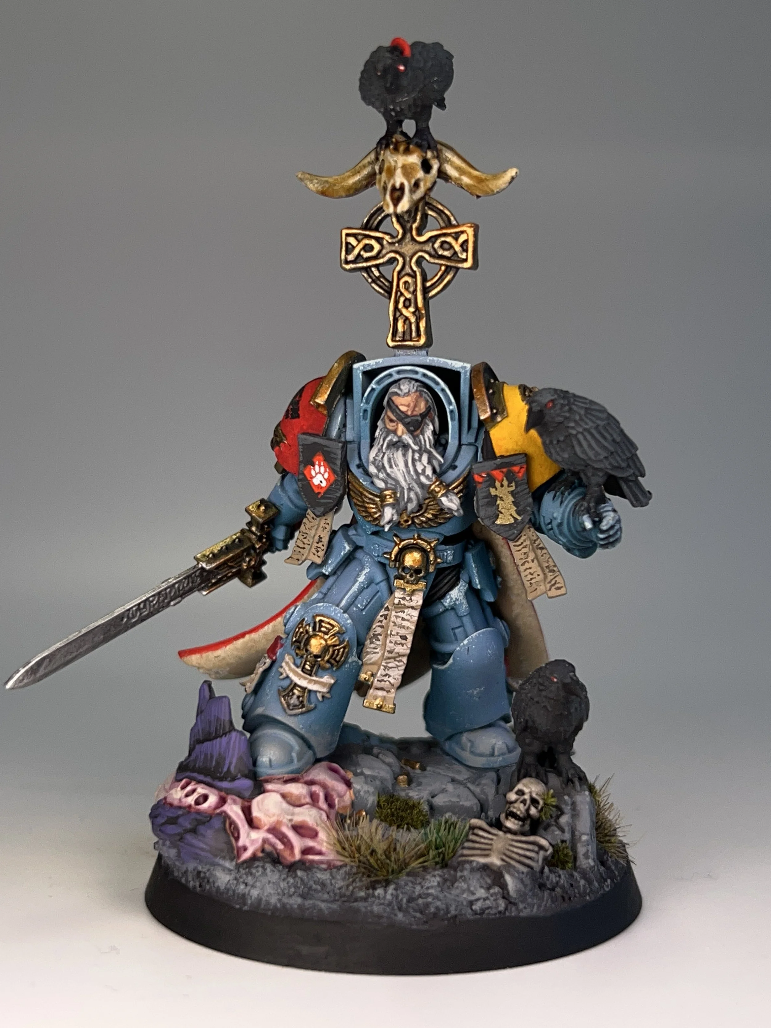 A detailed painted miniature figurine of a fantasy warrior in blue armor, holding a sword, with a cape, and surrounded by skulls and bones, standing on a circular base decorated with bones, skulls, and terrain.