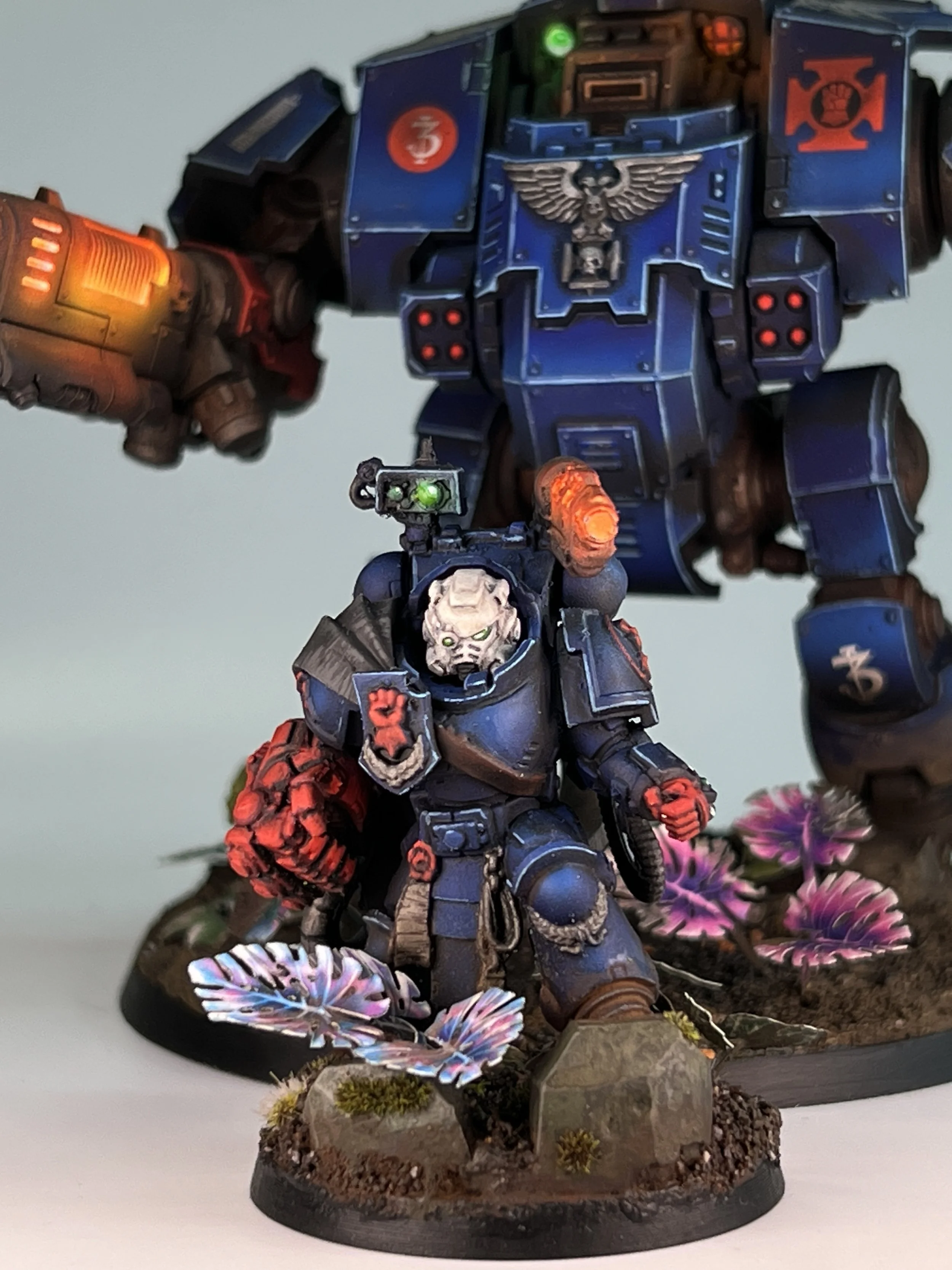 A painted miniature figurine of a futuristic soldier wearing dark armor with skull motifs, holding a gun, standing on a decorated base with flowers and moss. Behind the soldier is a large blue armored mech with various symbols, insignias, and bright 