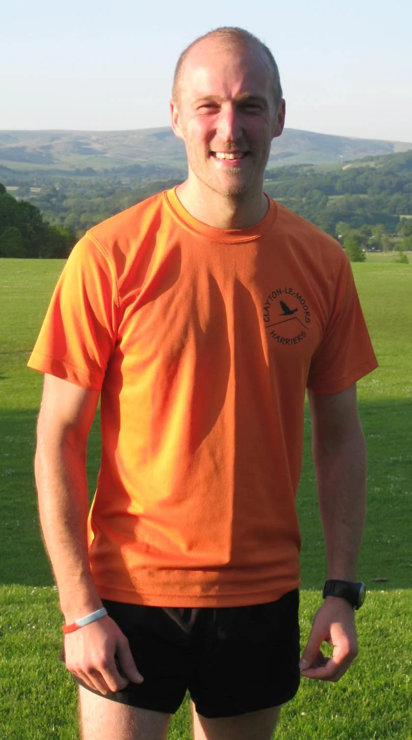 A man in an orange T-shirt and black shorts standing outdoors on a grassy field with hills in the background during daytime.