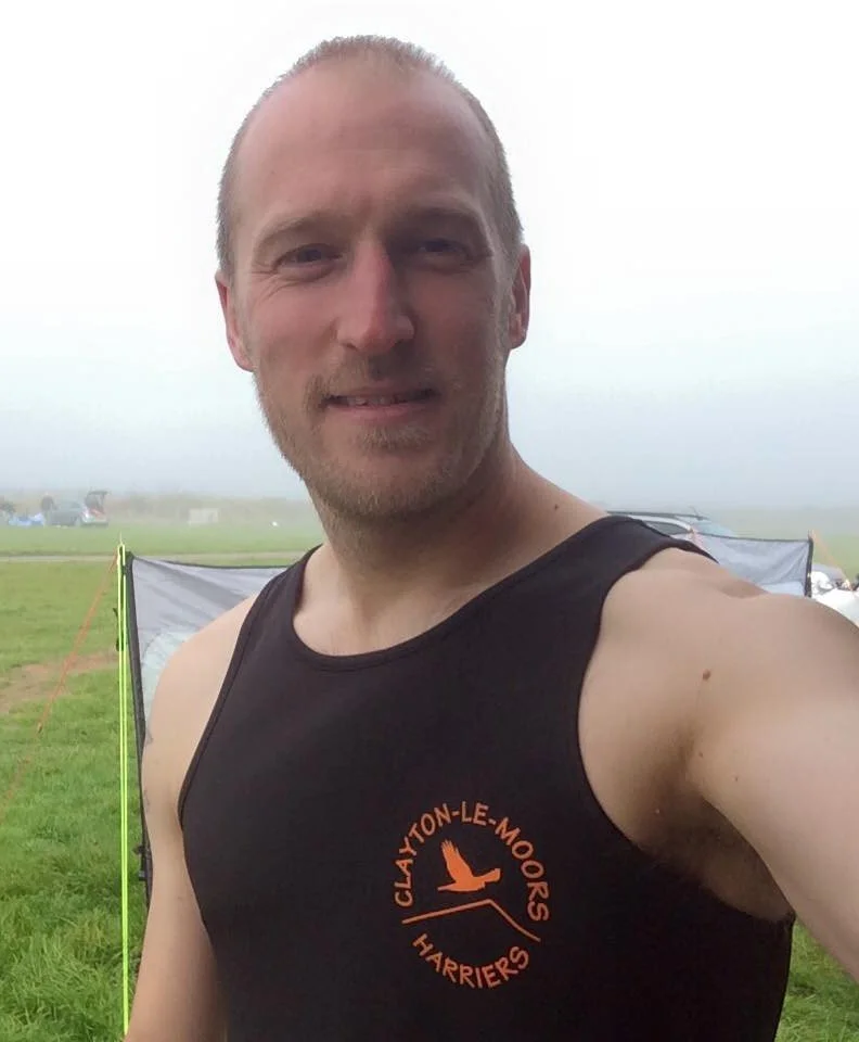 A man with a shaved head and beard taking a selfie outdoors on a foggy day, wearing a black sleeveless athletic shirt with orange text and logo, standing in front of a grassy field with tents and cars in the background.