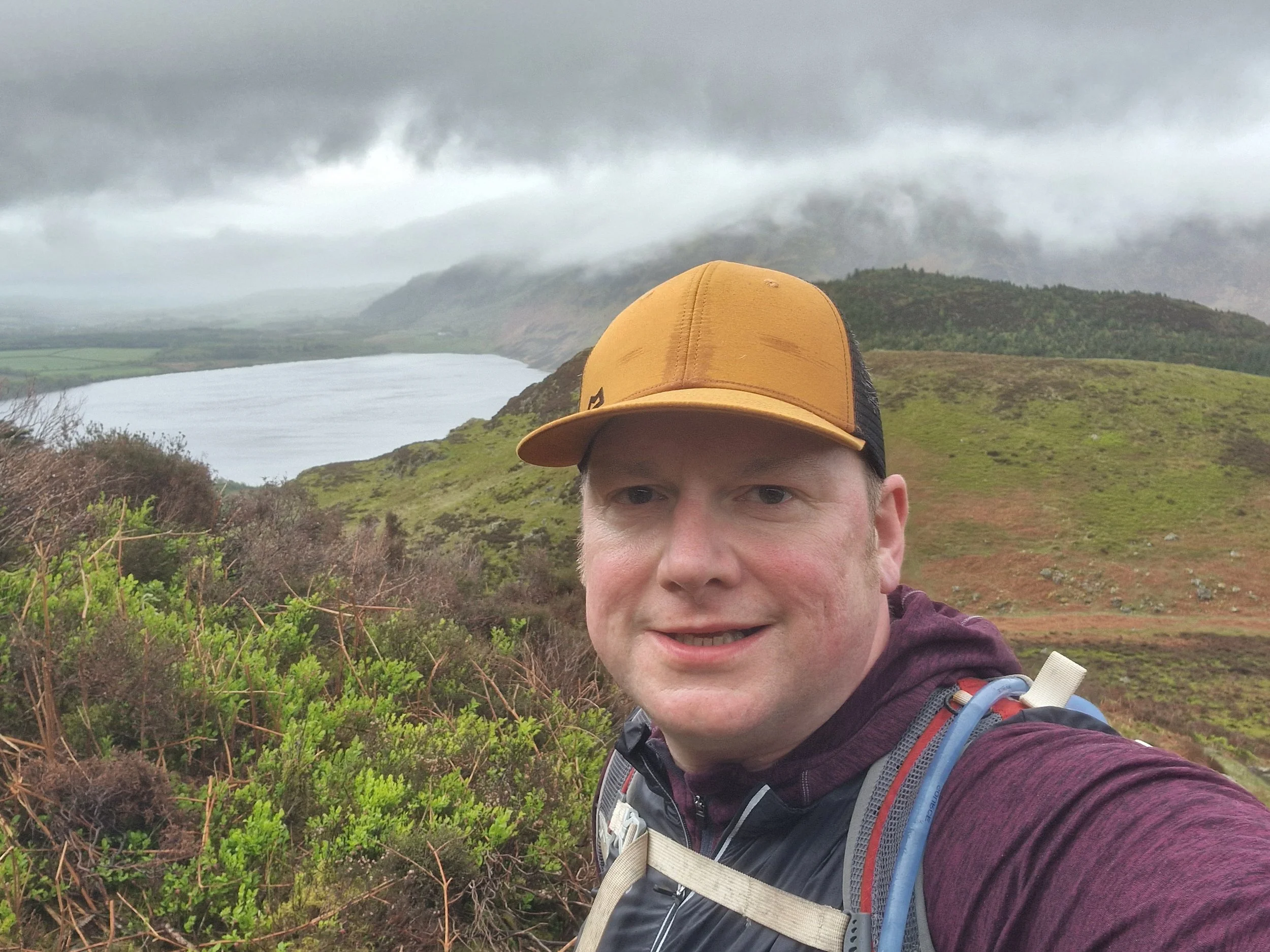 A man wearing a yellow cap and a hiking backpack taking a selfie during a hike in a green, mountainous landscape with a lake and cloudy sky in the background.