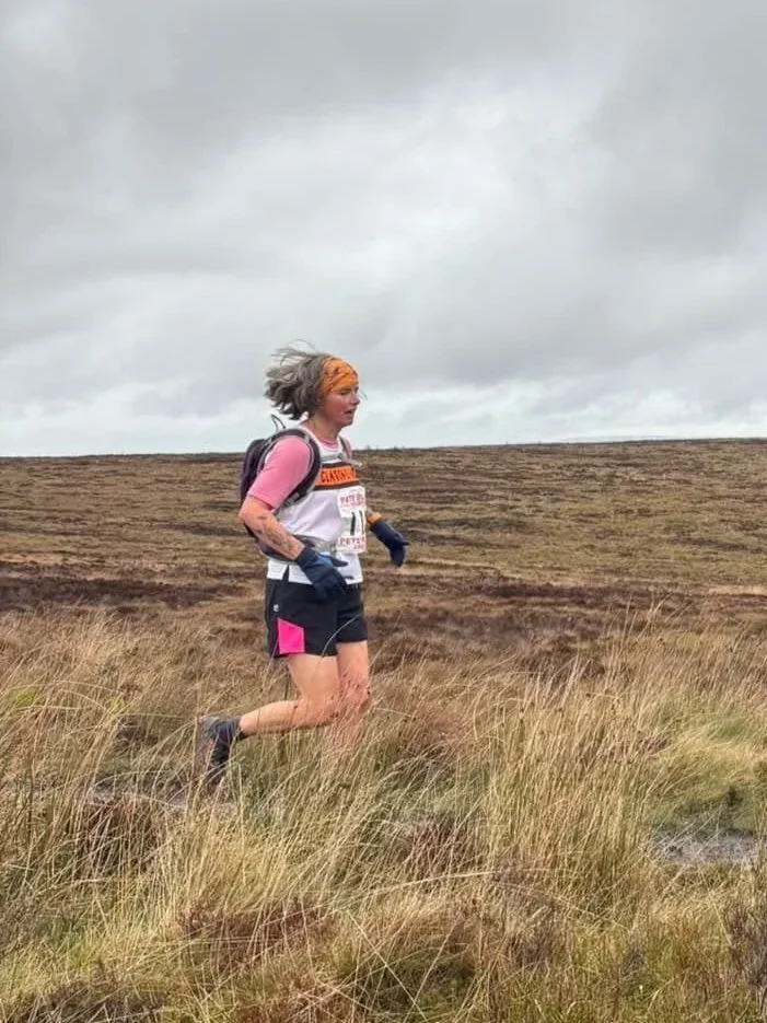A woman running through a grassy, open landscape with a cloudy sky overhead, dressed in athletic gear and wearing a race bib.