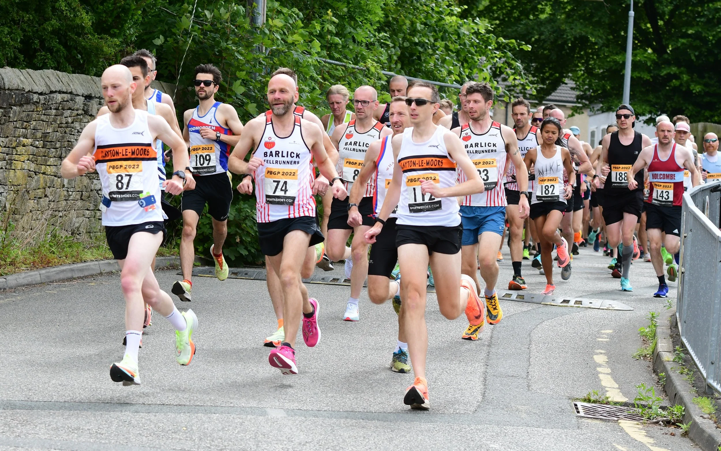 A group of runners participating in a race on a street with greenery and a stone wall in the background.