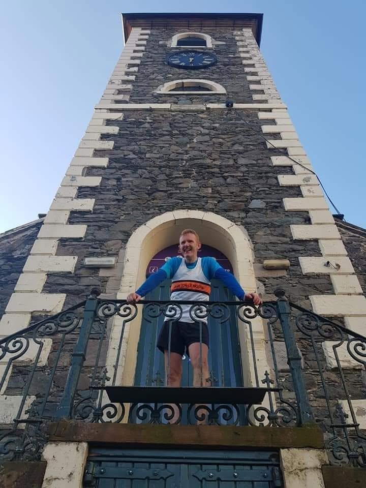 A man wearing running clothes and a race bib stands on a small balcony in front of a stone clock tower, smiling at the camera.
