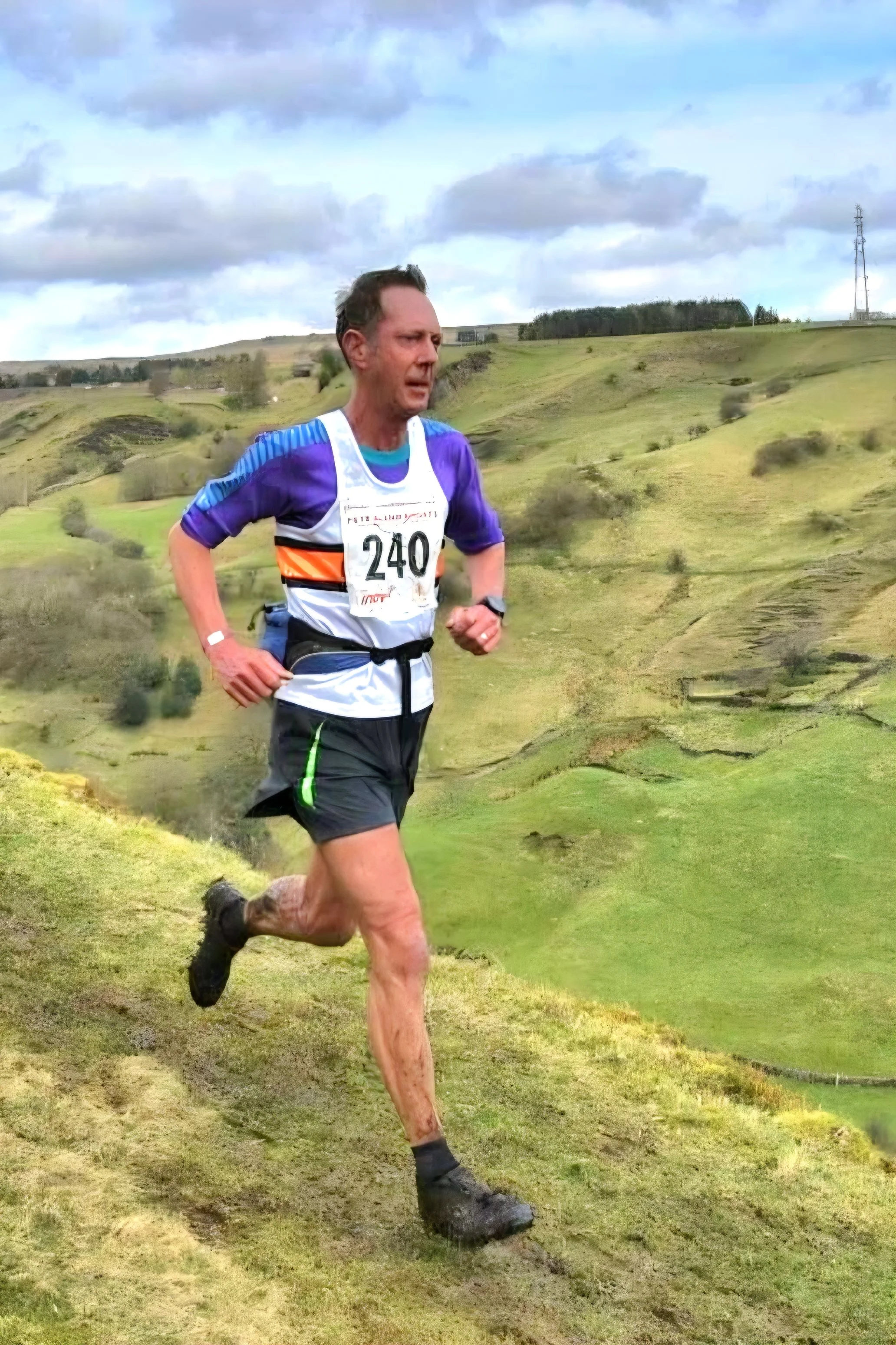 A man running outdoors on a dirt trail through rolling green hills under a partly cloudy sky, wearing a race bib number 240.