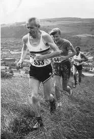 A group of runners participating in a race on a grassy hillside, with a rural landscape in the background.