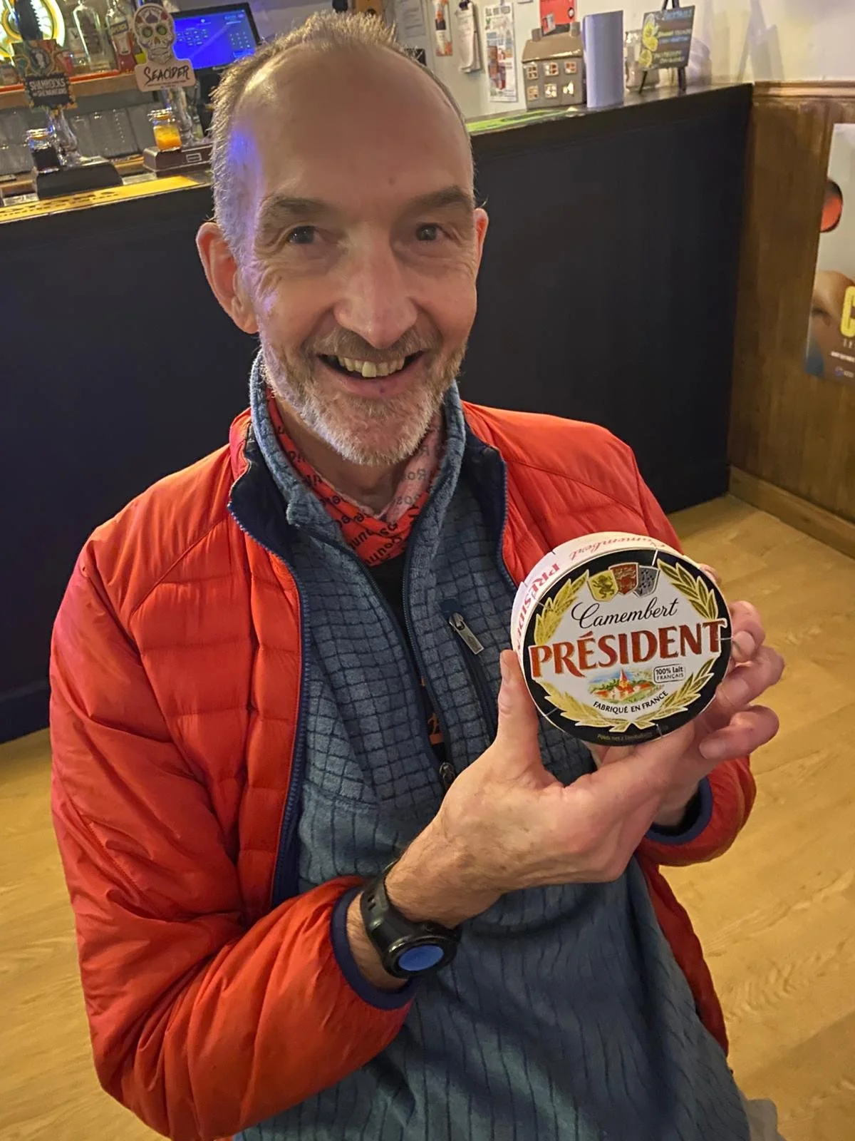 A smiling man with a beard and short hair, wearing a red jacket and holding a wheel of Camembert cheese labeled 'Président' in a restaurant or bar setting.