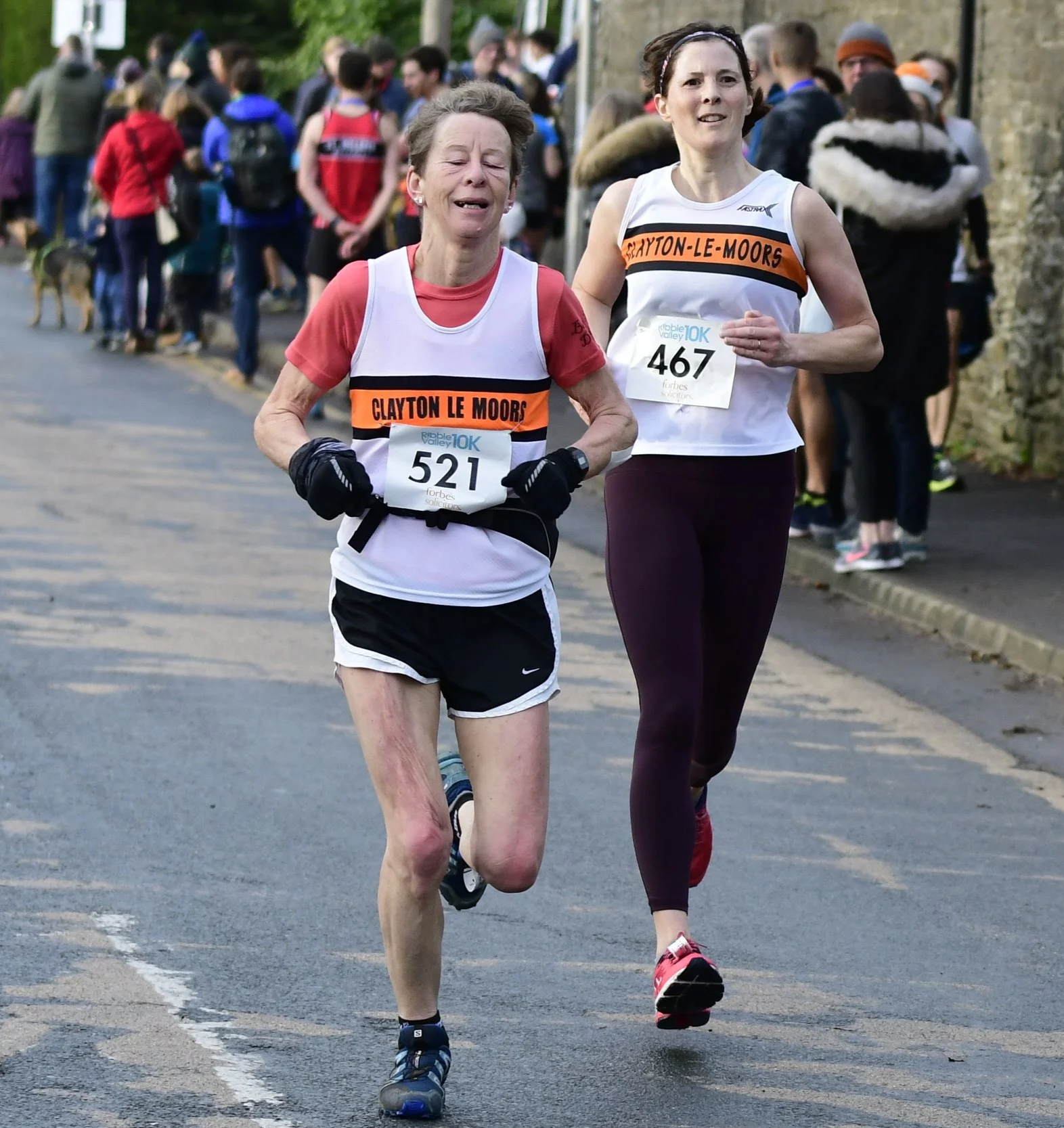 Two women running in a race, wearing athletic clothing and bib numbers 521 and 467, with a crowd of spectators in the background.