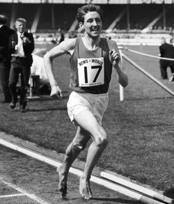 Black-and-white photo of male marathon runner wearing race bib number 17, running on a track at a stadium, smiling with onlookers in the background.