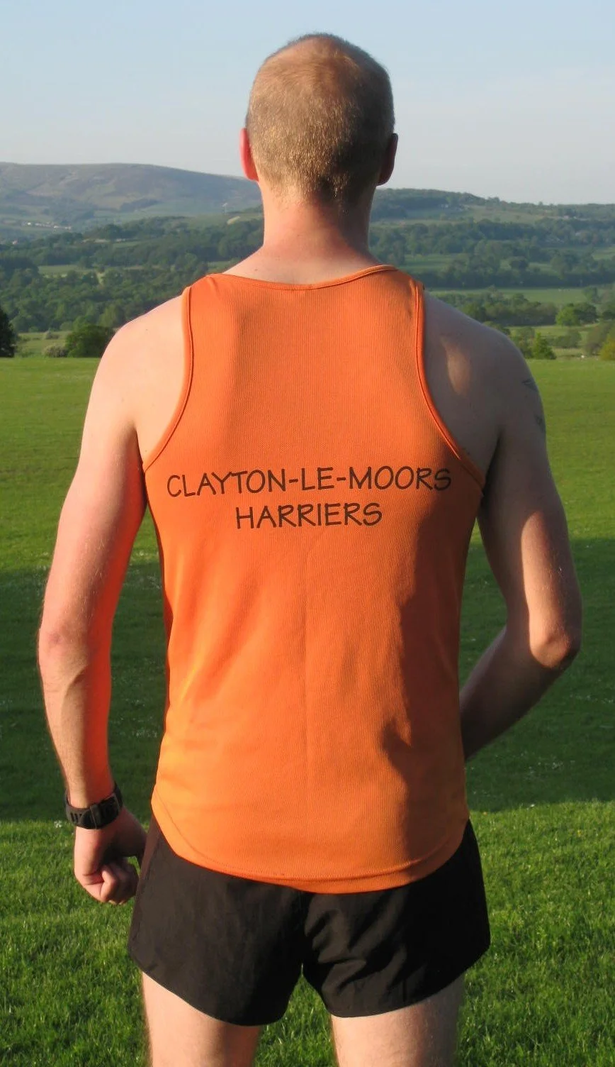 Back view of a man with short hair wearing an orange sleeveless sports jersey with "Clayton-Le-Moors Harriers" printed on the back, standing on grass with rolling hills in the background.