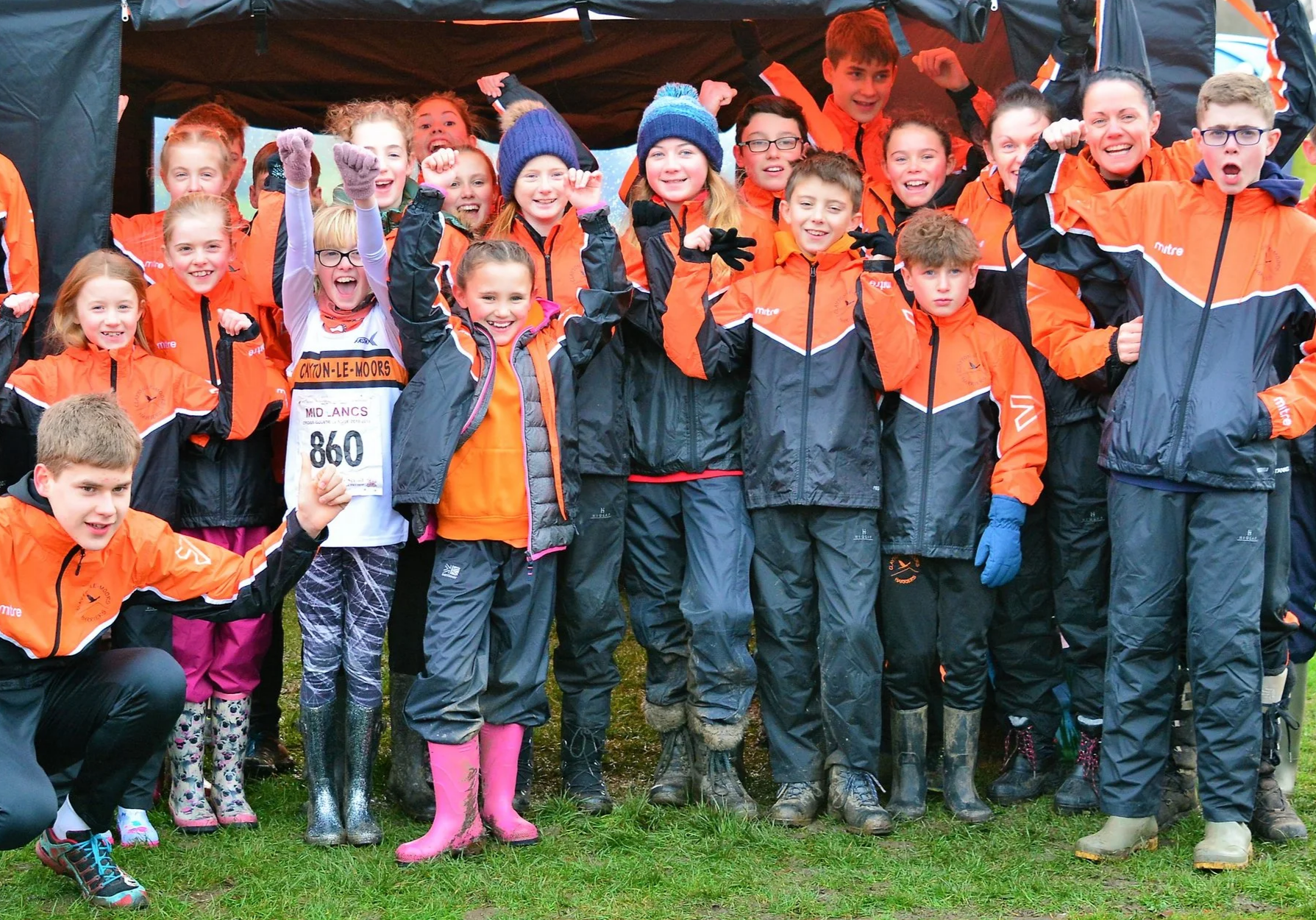 A group of children dressed in orange and black outdoor jackets, some with rain boots, posing under a black canopy in a grassy outdoor area. They are smiling, cheering, and making fist gestures, indicating excitement or celebration.
