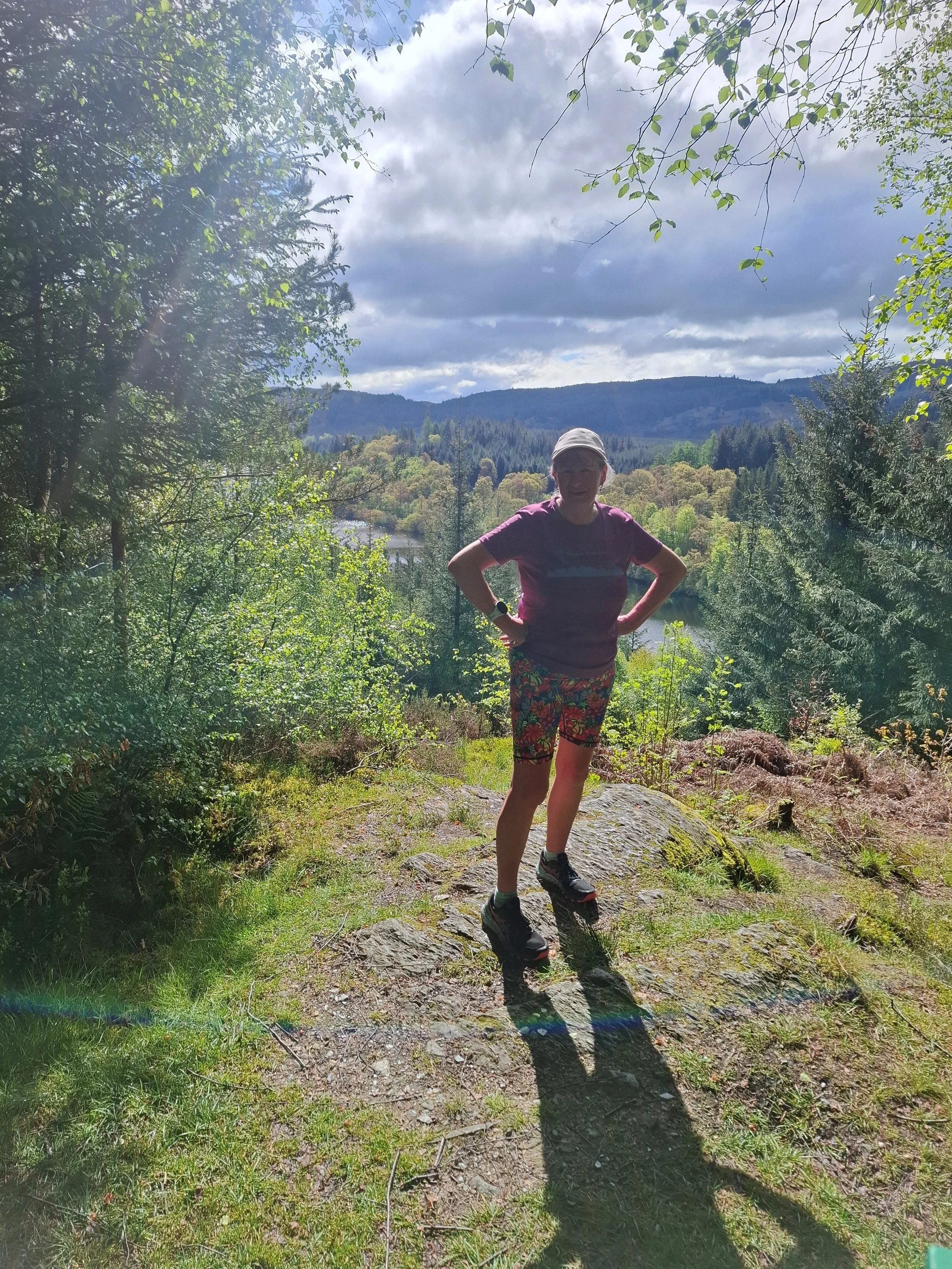 A person standing on a rocky trail in a forested area, with mountains, trees, and a cloudy sky in the background.