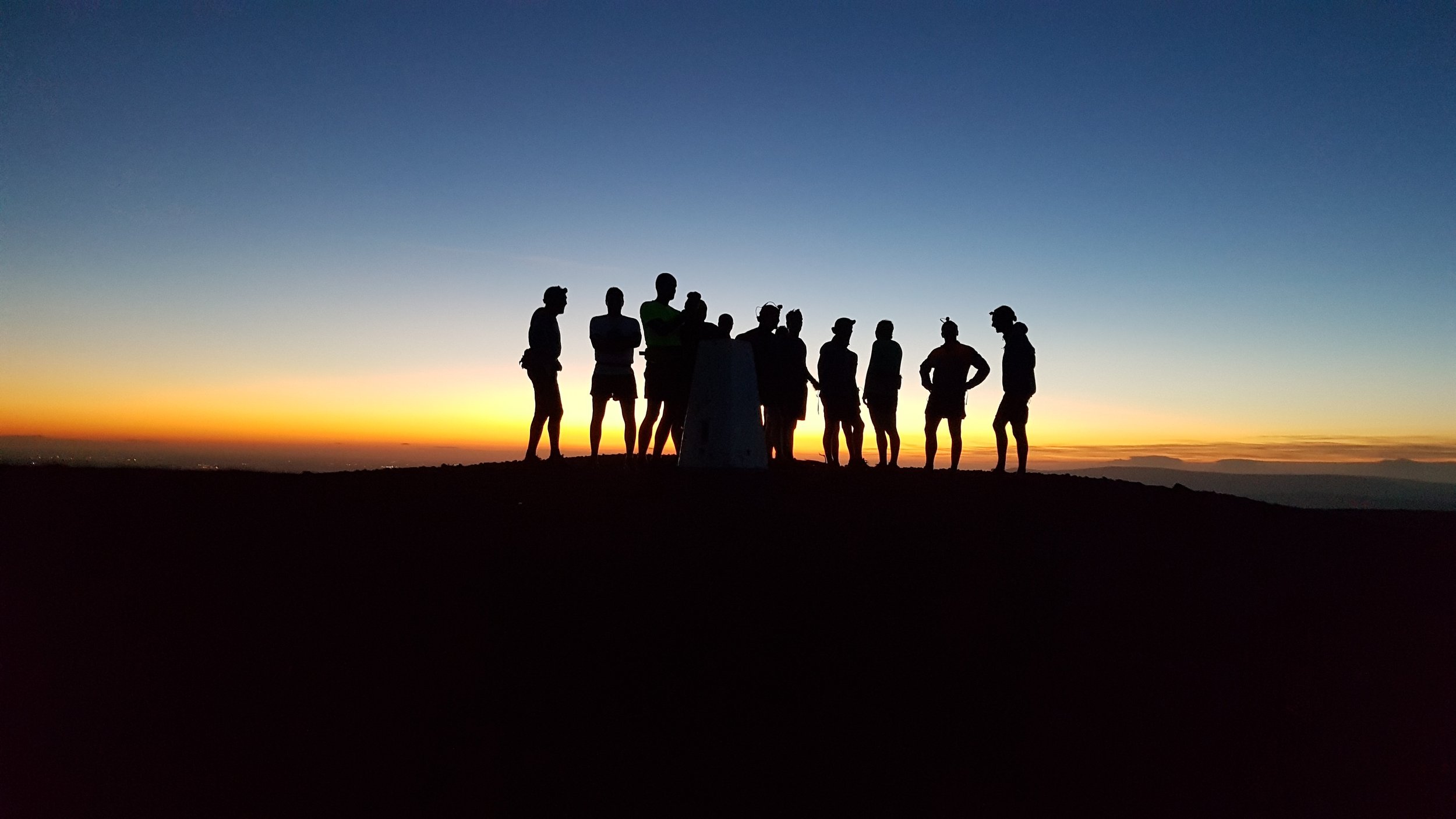 Group of people standing on a hilltop at sunset, silhouettes visible against a colorful sky with orange, yellow, and blue hues.