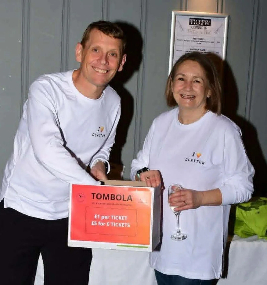Two smiling people at a charity event, one man and one woman, standing behind a table with a tombola box labeled with ticket prices, both wearing white shirts with the name 'Clayton' and a colorful logo.