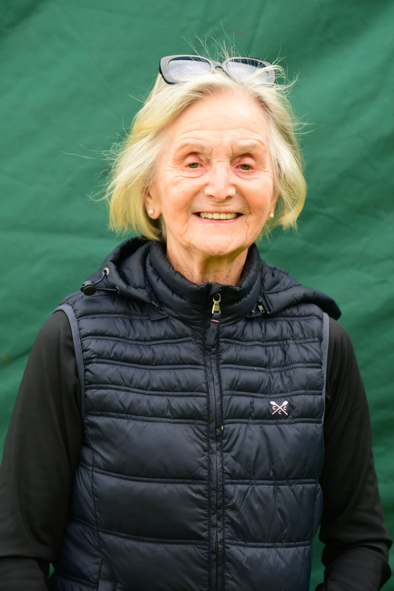 Smile of an elderly woman with short gray hair wearing sunglasses on her head, a pearl earring, and a black puffer vest over a black long-sleeve shirt, standing in front of a green background.