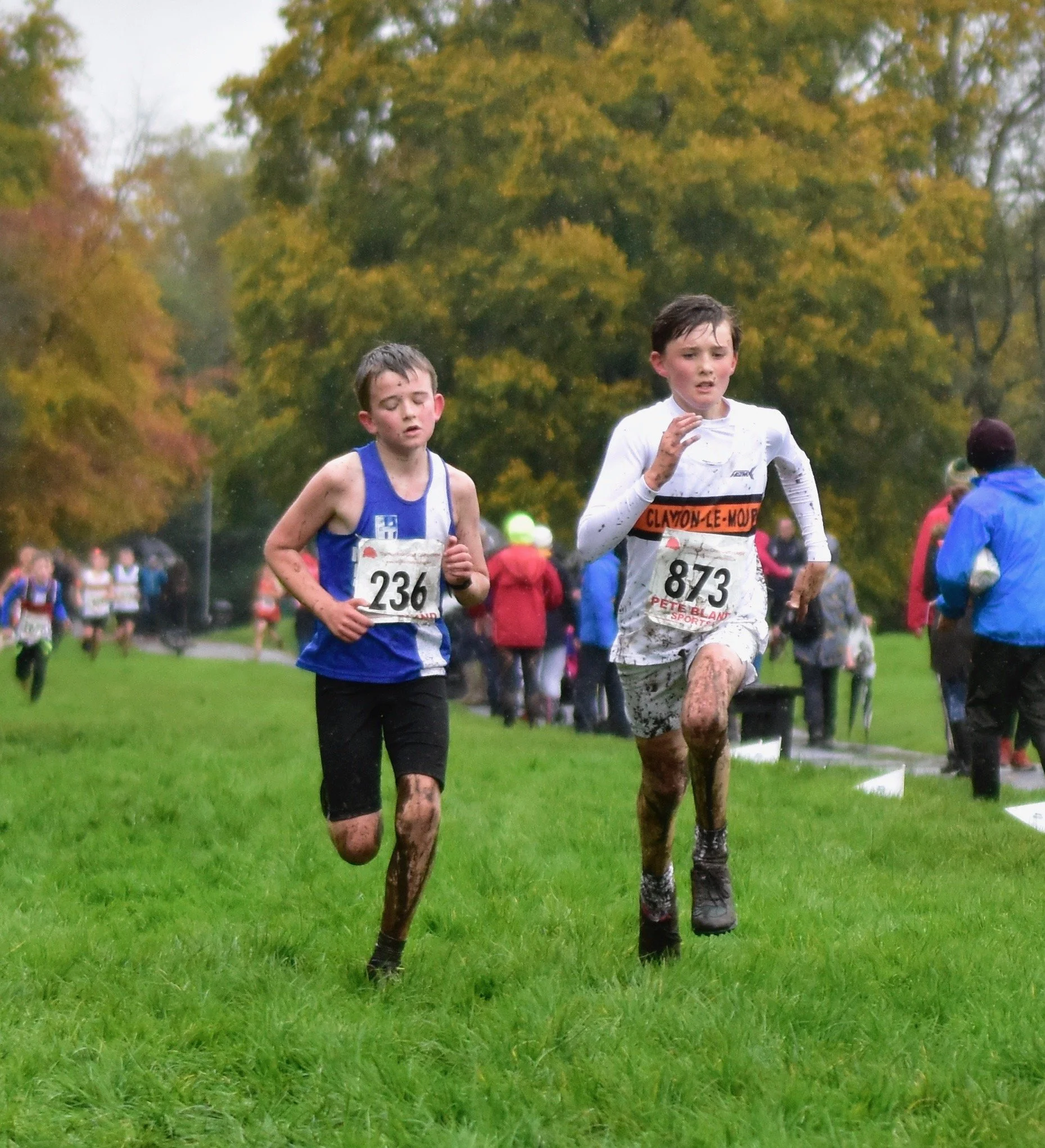 Two boys running in a muddy cross-country race on a grassy field, with a wooded area in the background, wearing race bibs and athletic clothing.