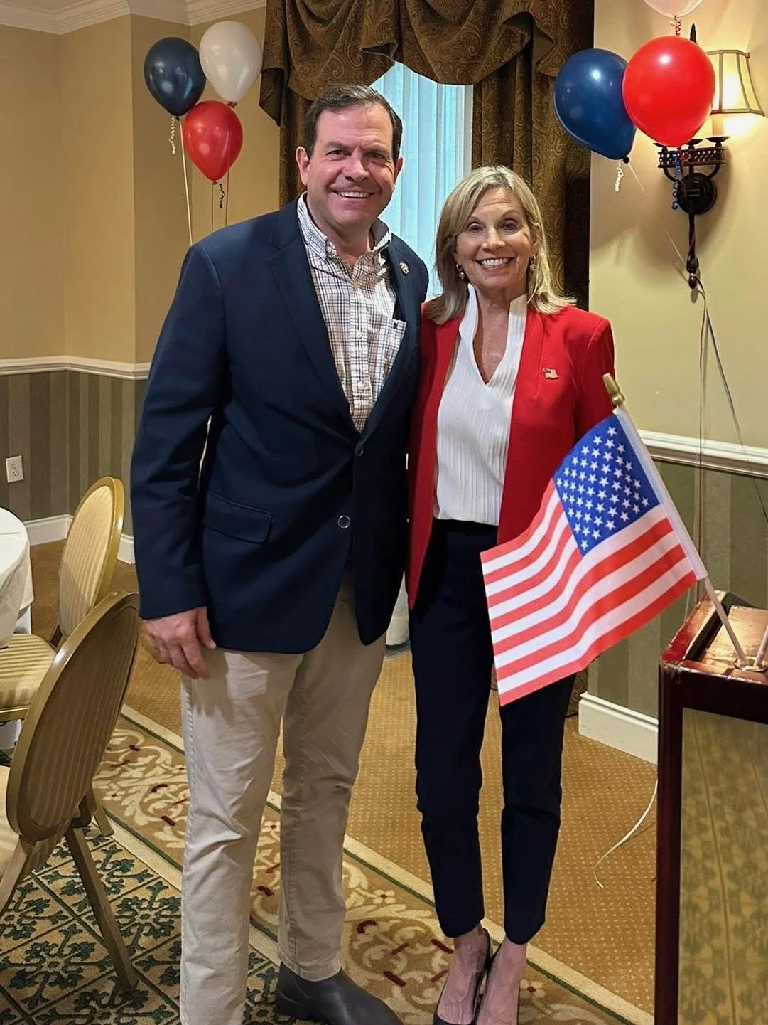 Nancy Campbell and Delegate Bill Wiley post at a patriotic, celebratory event. Nancy is dressed in a red blazer and black pants, and Bill is wearing a blue blazer and beige pants. The background features balloons in red, white, and blue.