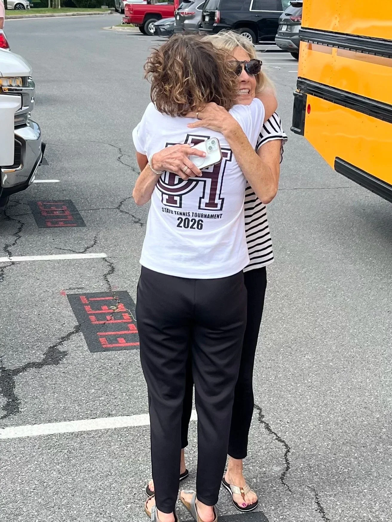 Nancy Campbell and a friend embrace in a parking lot in Winchester, Virginia, one wearing a white T-shirt with a tennis tournament logo, and the other in a striped shirt. There are parked cars and a yellow bus in the background.