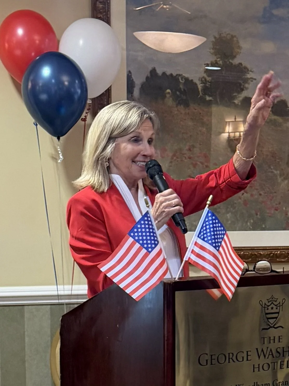 Nancy Campbell, a woman in a red blazer smiling and speaking into a microphone at a patriotic event, with American flags and balloons in red, white, and blue behind her, at the George Washington Hotel in Winchester, Virginia.
