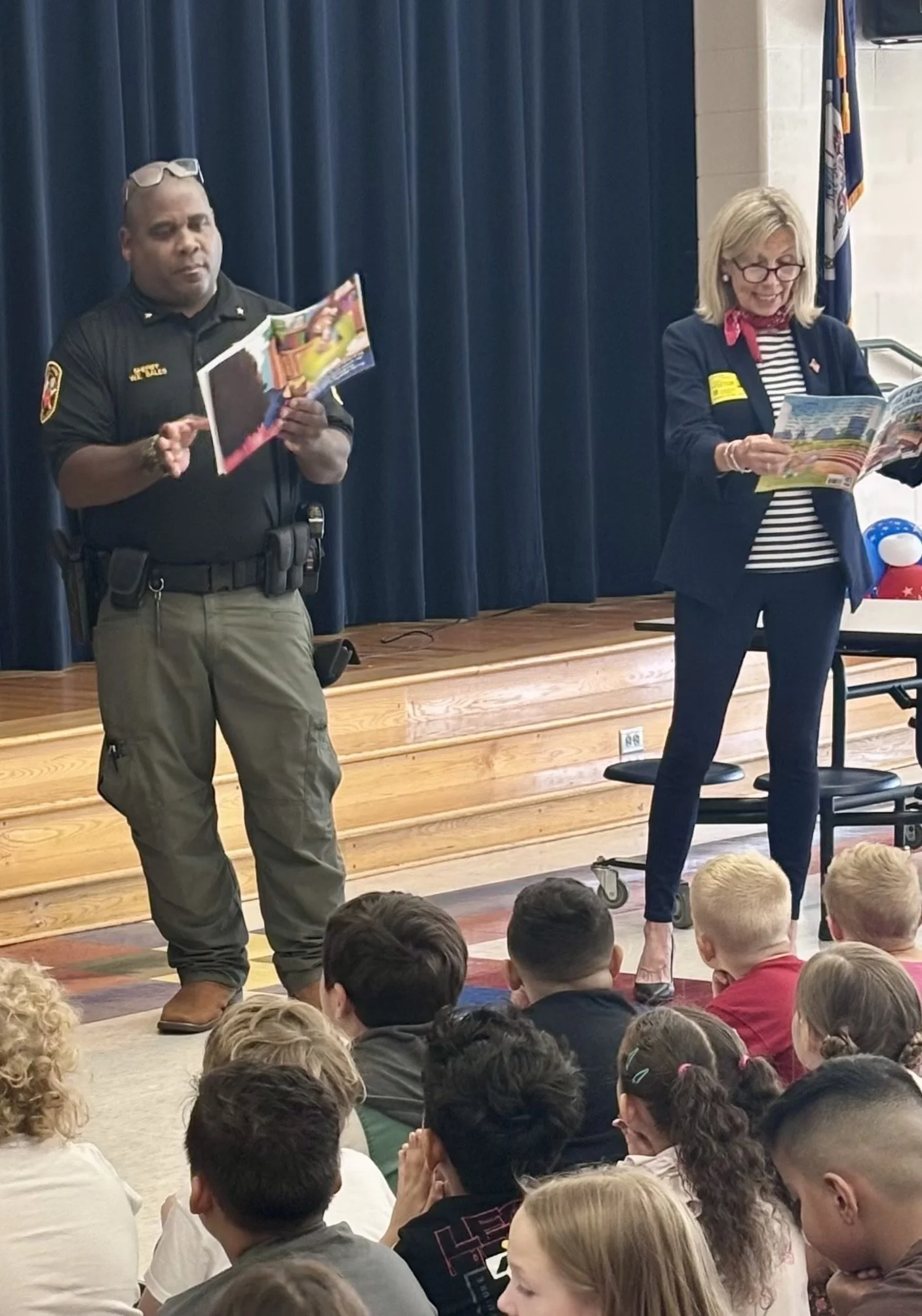 Sheriff William E. Sales Jr. in uniform, and Nancy Campbell in a blazer and striped shirt are standing on stage reading from booklets.  The audience, composed mostly of children, is sitting on the floor in front of the stage, attentively listening.