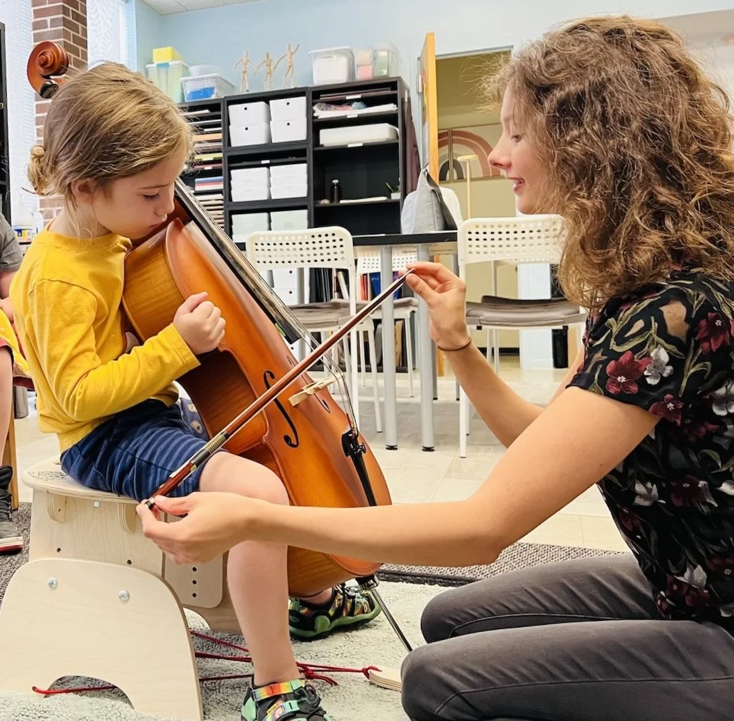 A young girl in a yellow shirt and shorts is sitting on a small wooden seat playing a violin. An adult woman with curly hair, wearing a floral top, is kneeling in front of her, helping her hold and play the violin with a bow. The background shows chairs, shelves with books and supplies, and a classroom setting.