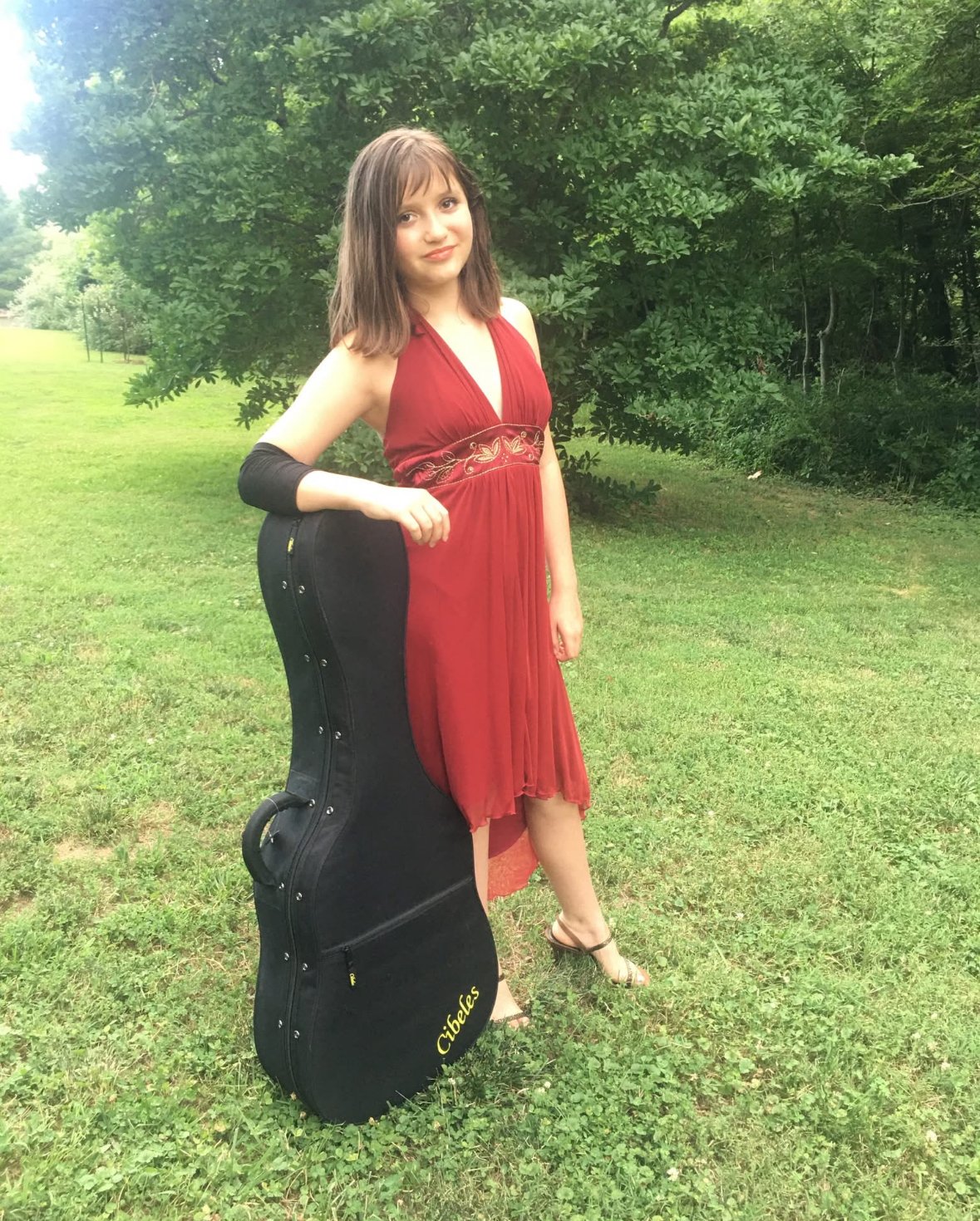 A young woman in a red dress with embroidered detail on the waist stands outdoors in a grassy area with green trees in the background. She has shoulder-length brown hair and is holding a black guitar case with the name 'Cibeles' on it.