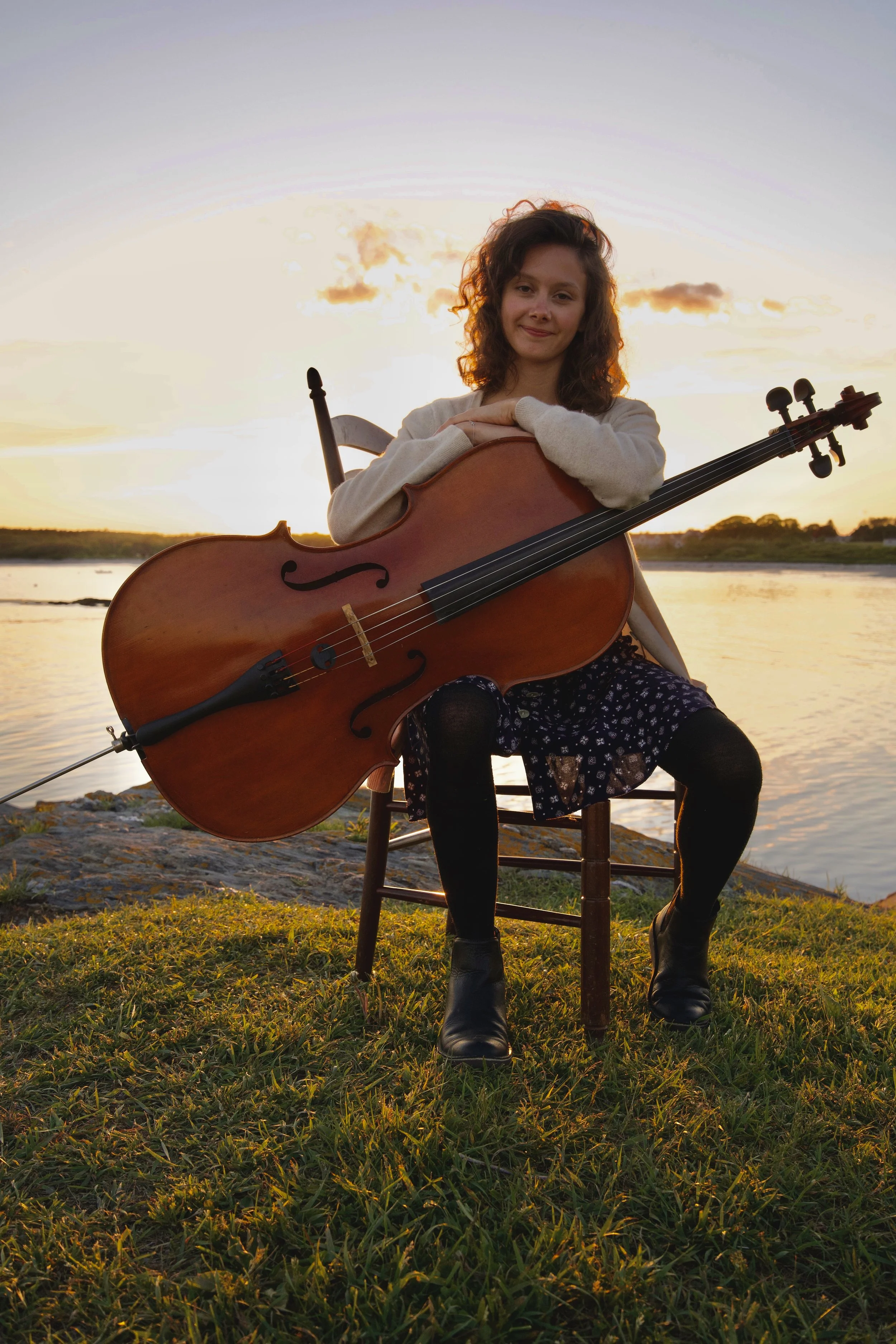 A young woman with curly brown hair sitting on a wooden chair outdoors during sunset, holding a cello, with water and a sunset sky in the background.