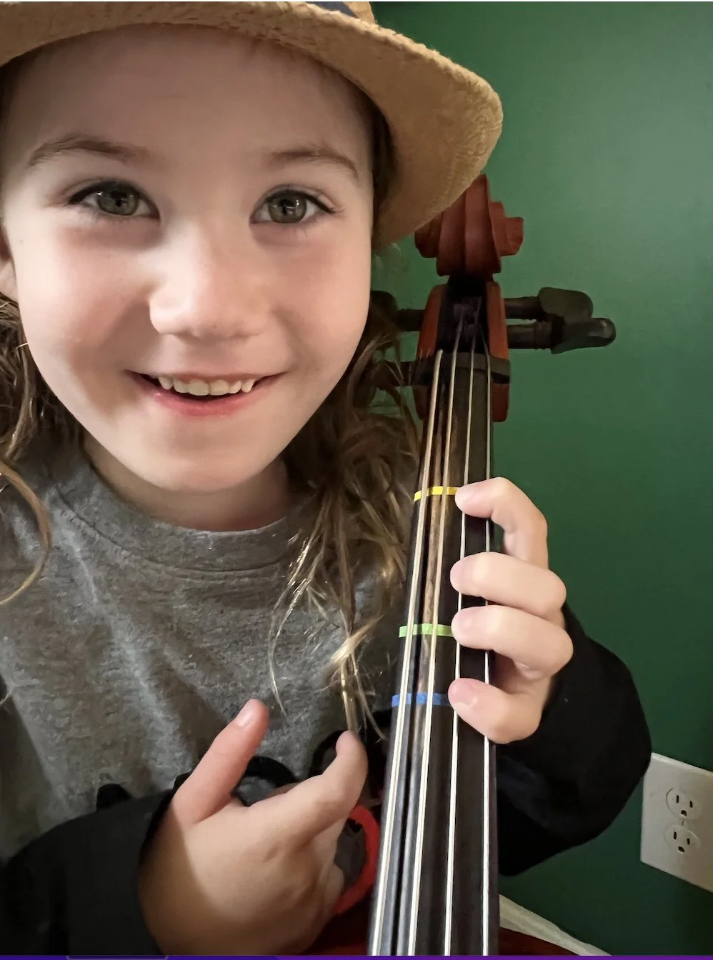 A young girl with light brown hair, wearing a straw hat and a grey shirt, smiling and holding a violin.