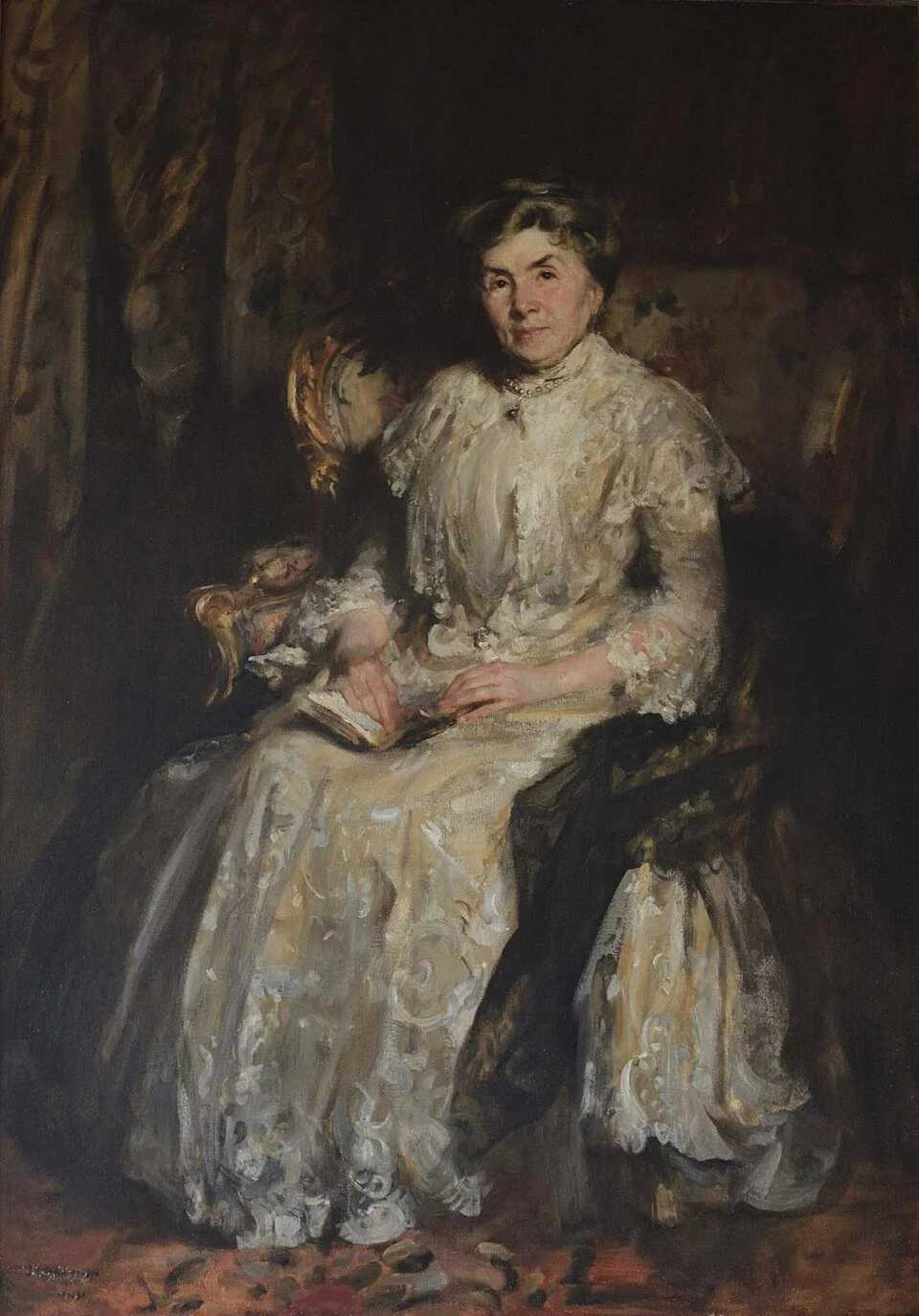 An elderly woman sits for a portrait, looking directly at the viewer as she holds a book in her lap.