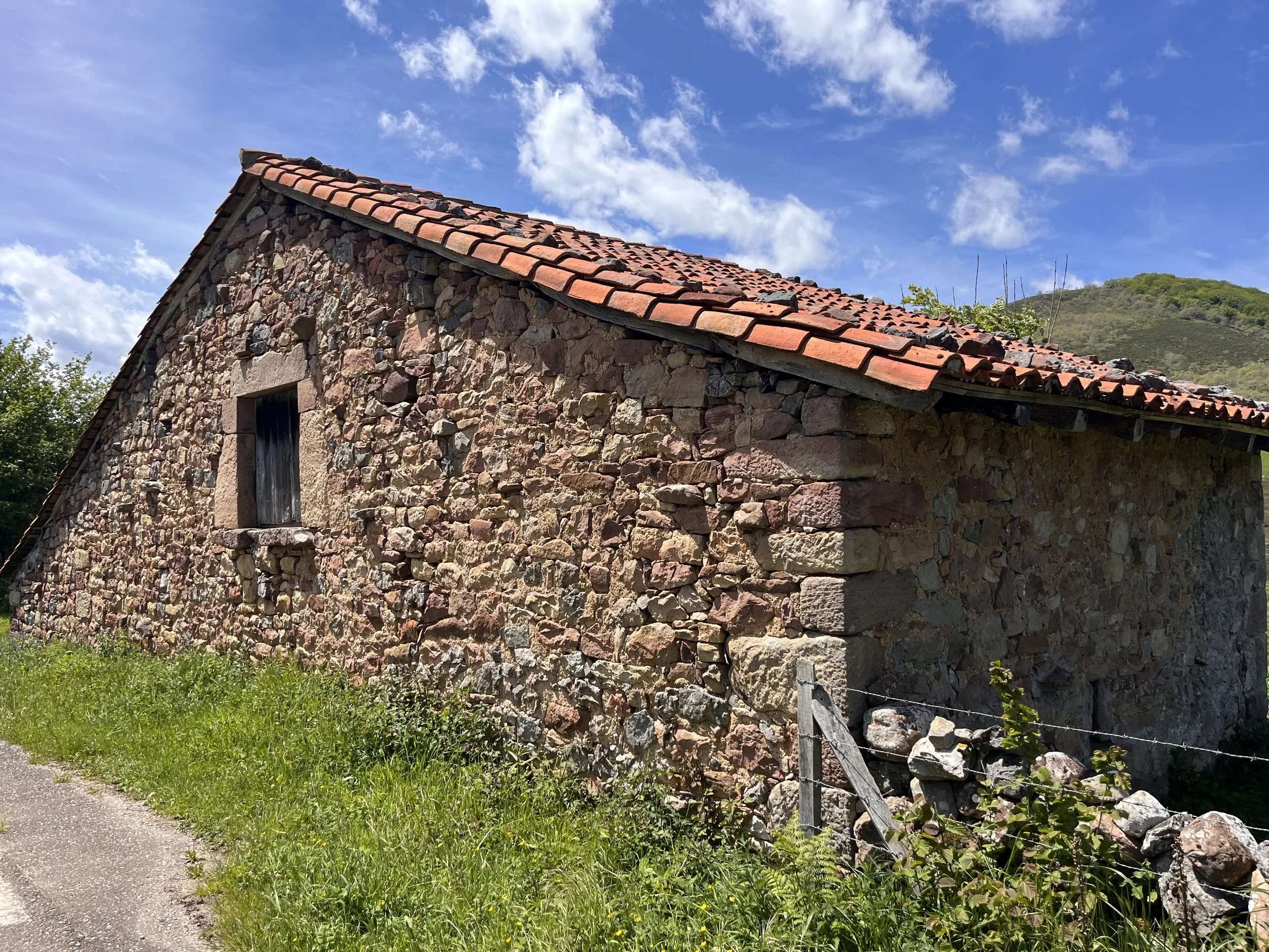 A stone building with a red tile roof and a small window, surrounded by green grass and plants, with a background of hills and a partly cloudy sky.