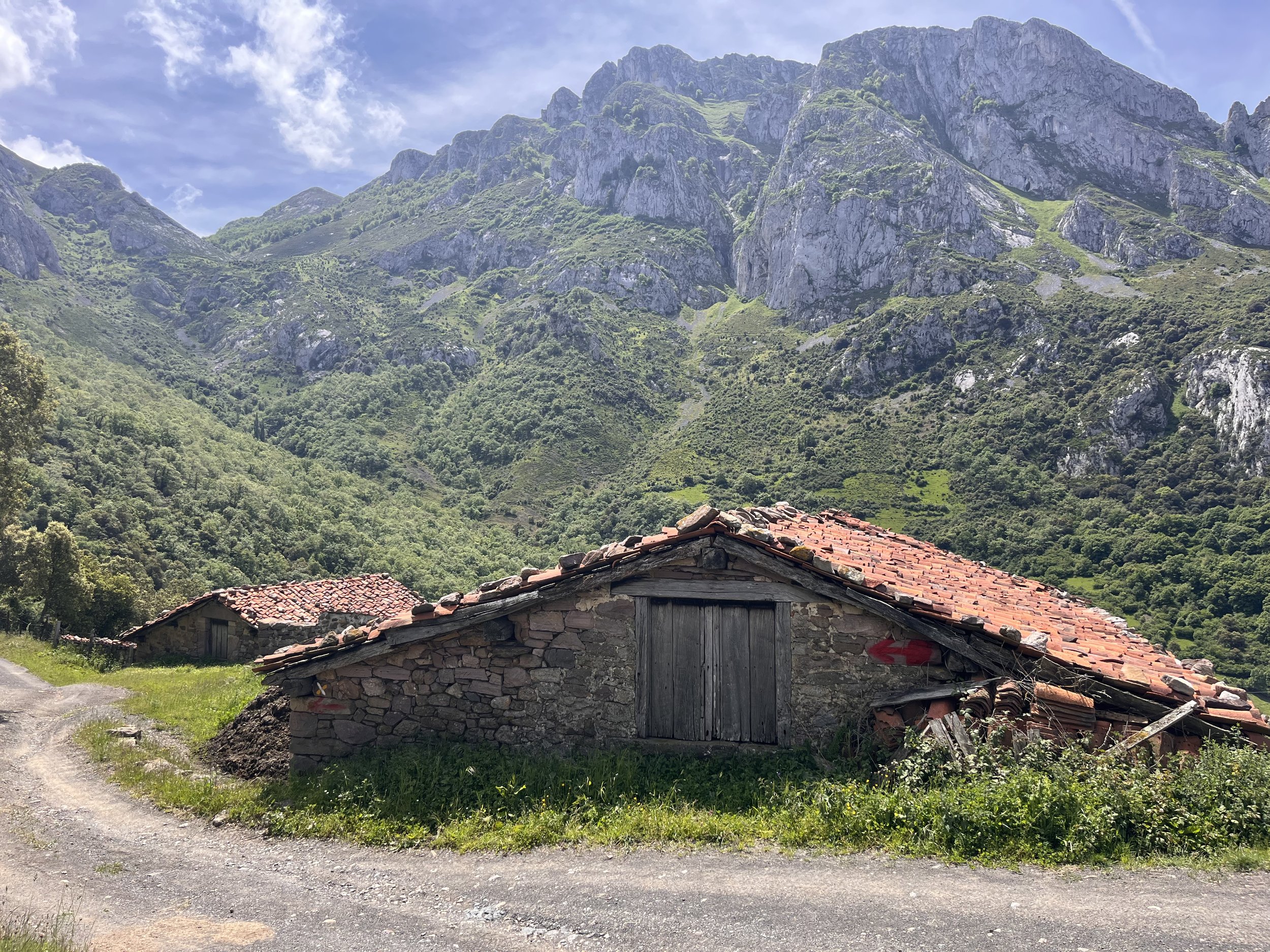 Old stone and wood barn with a red tiled roof in a rural area, with green mountains in the background and a partly cloudy sky.
