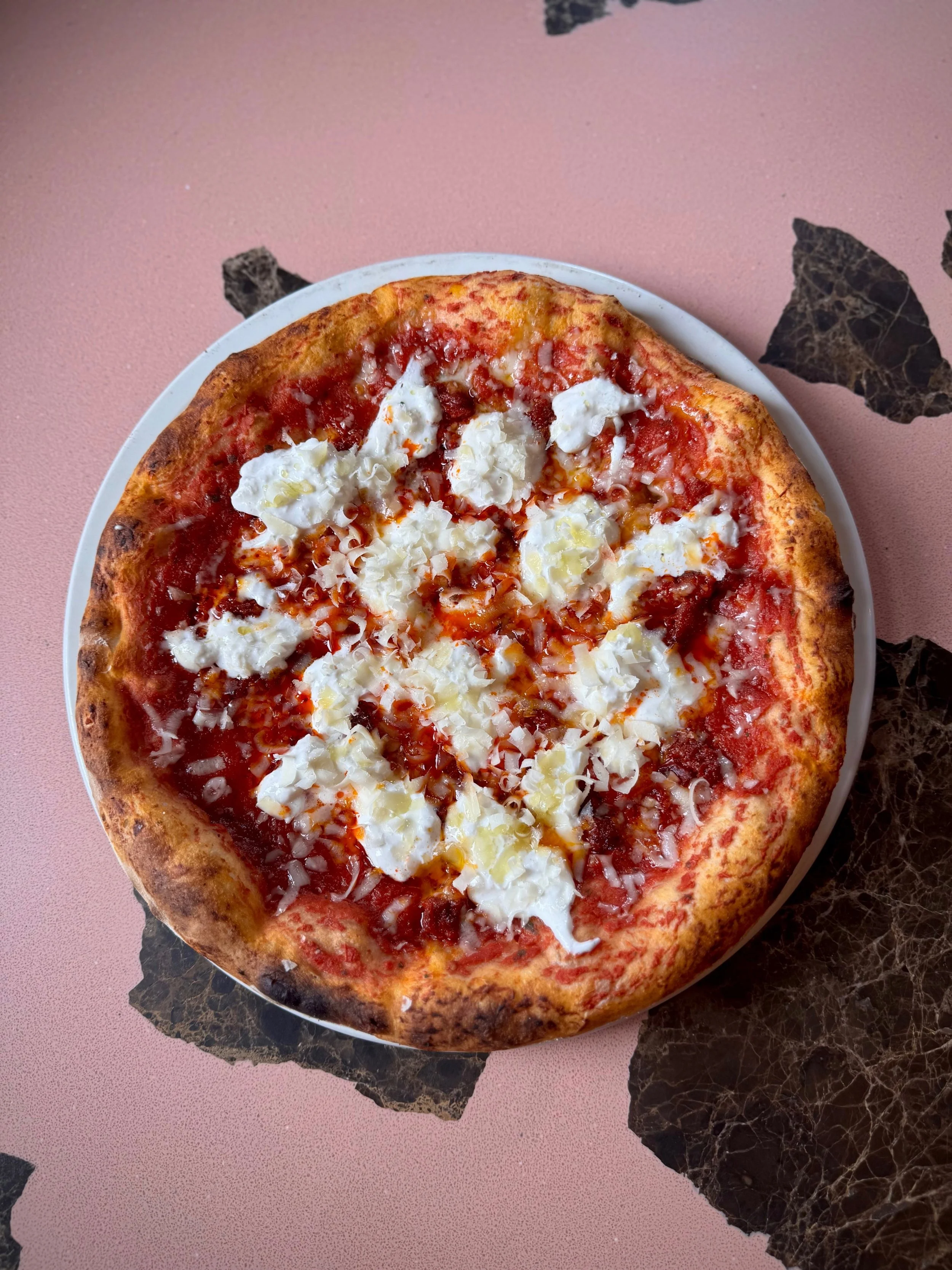 A pizza with cheese, tomato sauce, and cheese on a white plate on a black marble table with a pink background.