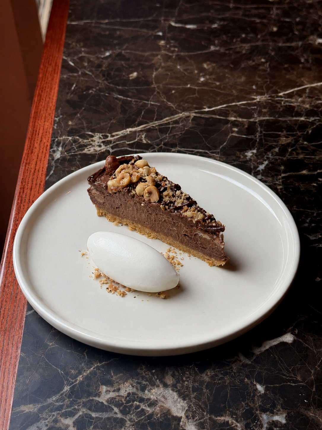 Chocolate cheesecake topped with chopped nuts on a white plate, served with a dollop of whipped cream on a dark marble table.