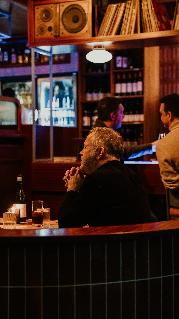 Man with gray hair and beard sitting at a bar with candles, bottle, and glass of dark beverage, in a cozy dimly-lit restaurant or bar with wooden shelves and records, and two people talking in the background.