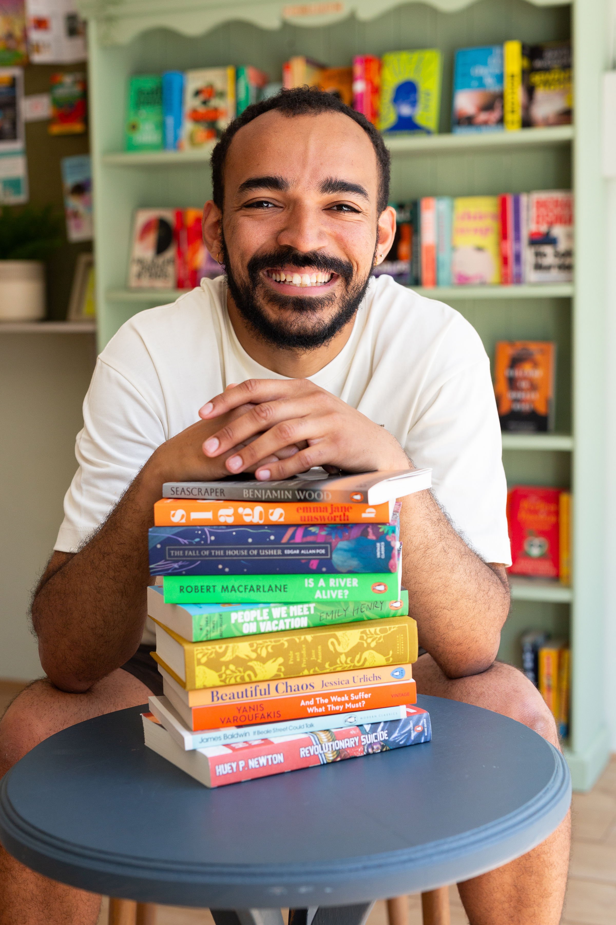A smiling man with a beard and earrings, sitting behind a tall stack of books on a table, in a room with a bookshelf in the background. The books are colourful and arranged vertically.