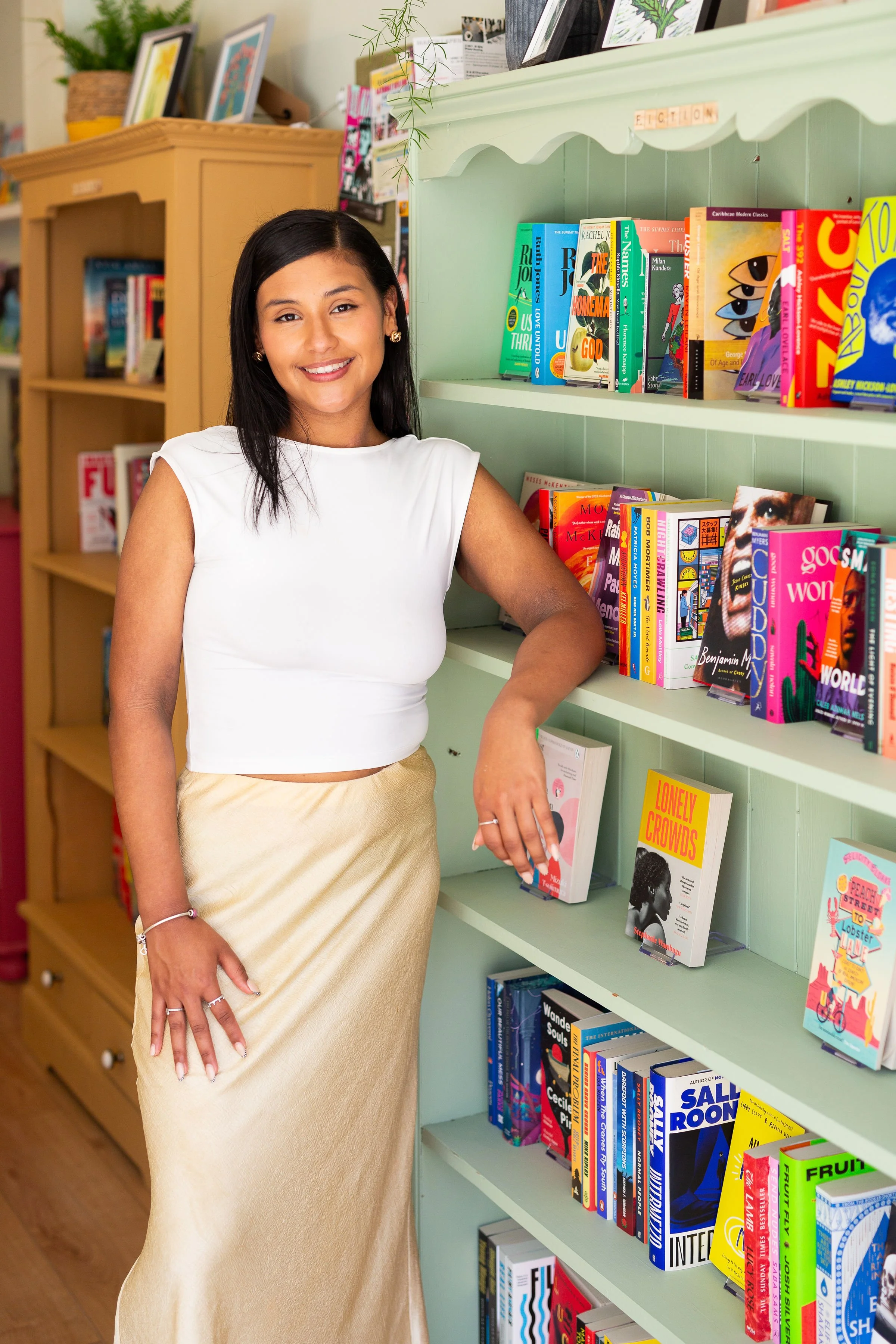 A young woman with long dark hair, wearing earrings, a white sleeveless top, and a gold skirt, standing in a bookshop, smiling at the camera. She is next to a light green bookshelf filled with colourful books.