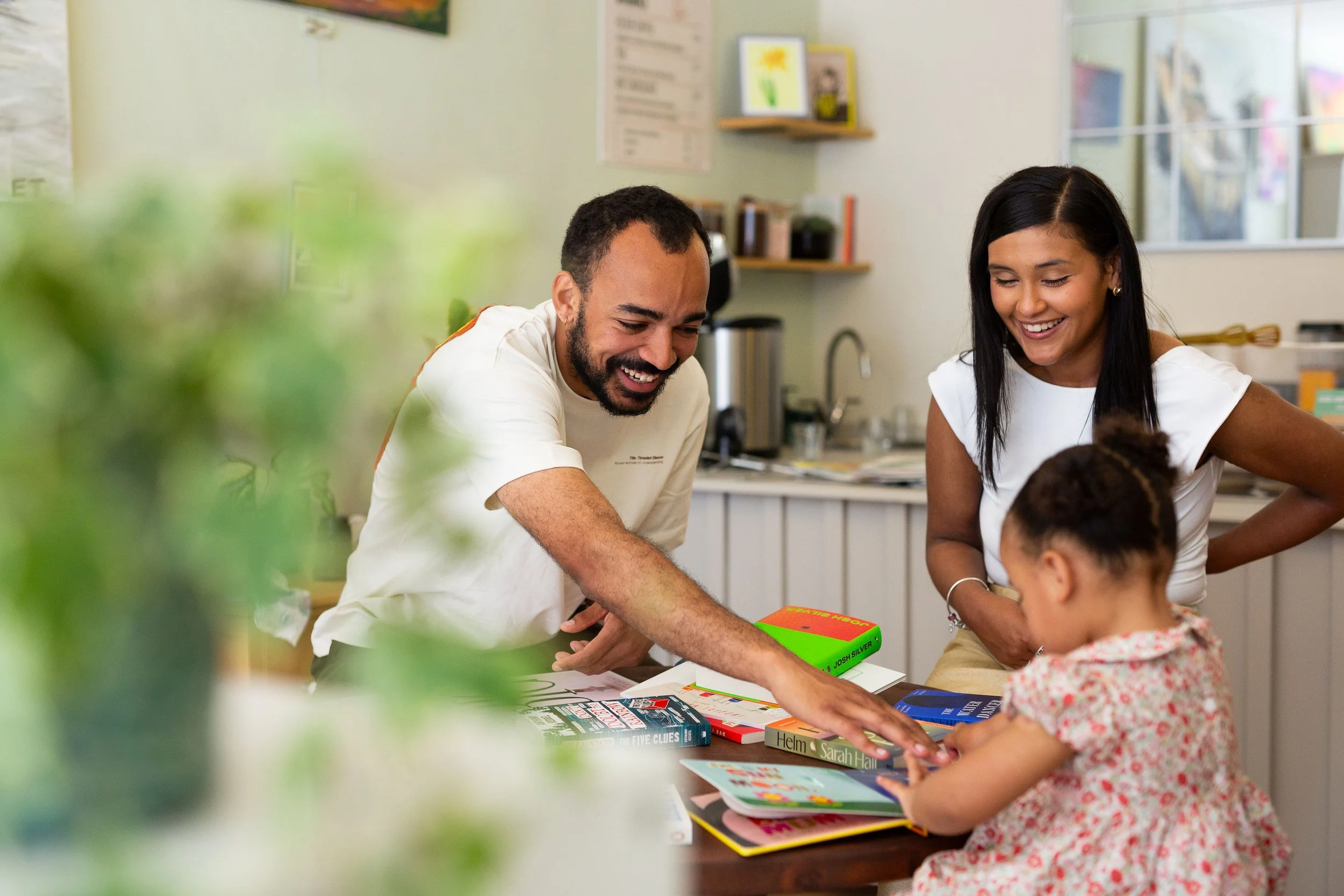 A smiling man and woman are engaging with a young girl at a table filled with books, in a cozy, well-lit room with bookshelves and artwork in the background.