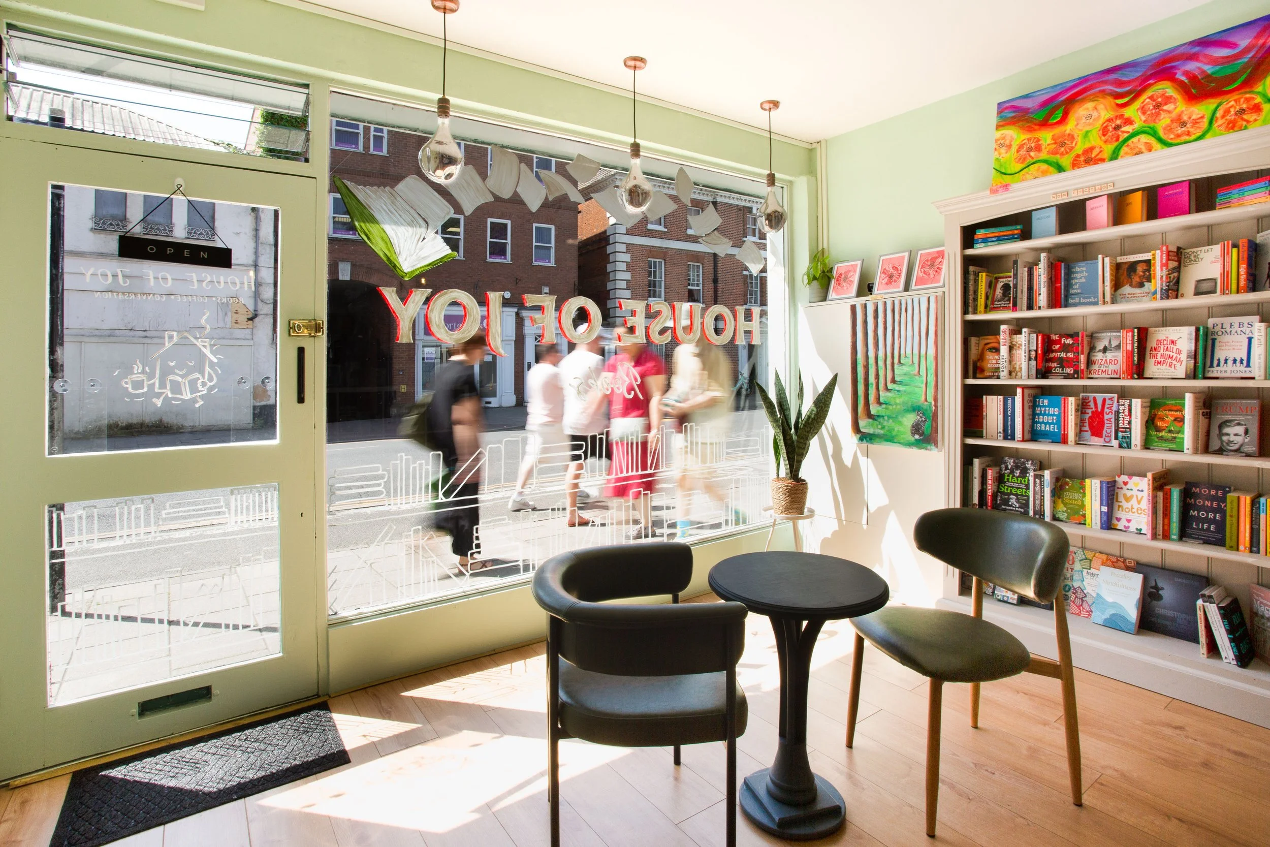 Inside a bookstore or library with pastel green walls, a black round table, and three black chairs. A large window displays the street outside with people walking past and buildings across the street. The window has red and white lettering that says 'HOUSE OF JOY' and a graphic of an open book and a coffee cup. To the right, a bookshelf filled with colorful books and framed artwork above it. There is a houseplant next to the bookshelf. An abstract, colorful painting with orange flowers and green stems is above the bookshelf.